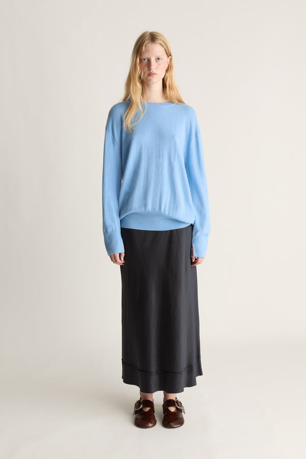 A woman stands against a plain light beige studio background wearing a light blue long-sleeve knit top, a long black skirt, and dark brown loafers.