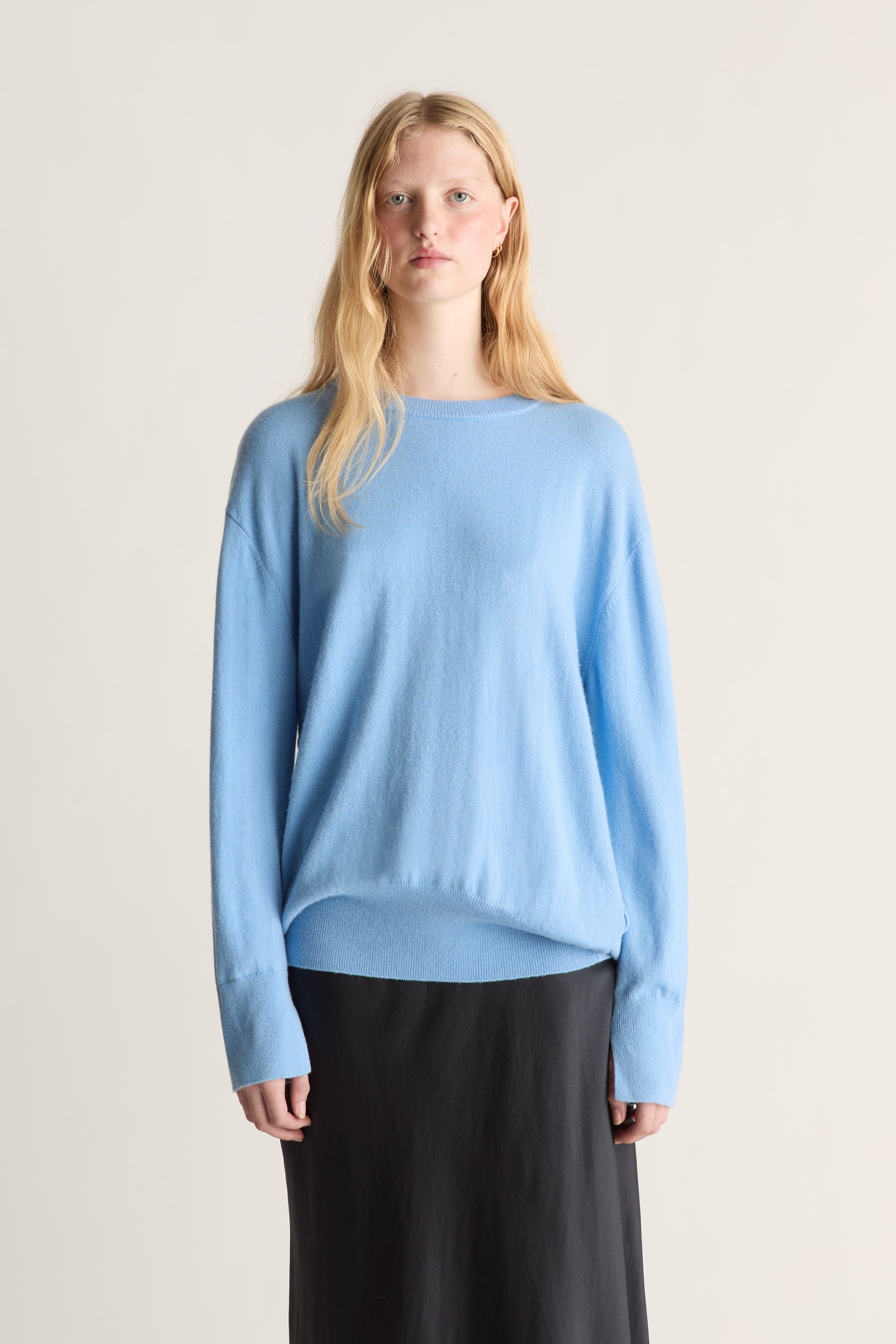 A blonde model wearing a light blue knit sweater and a black skirt, standing against a plain light beige studio background.