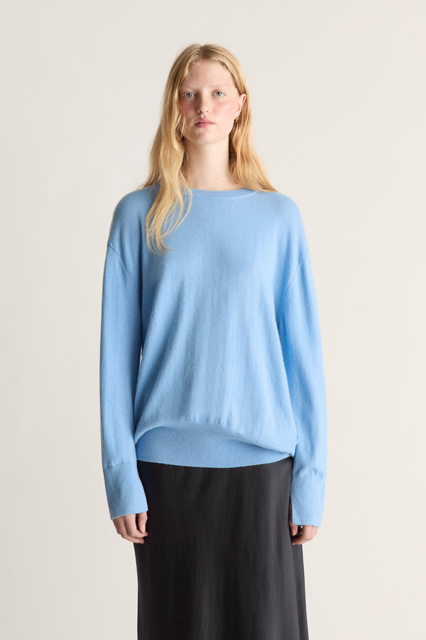 A blonde model wearing a light blue knit sweater and a black skirt, standing against a plain light beige studio background.