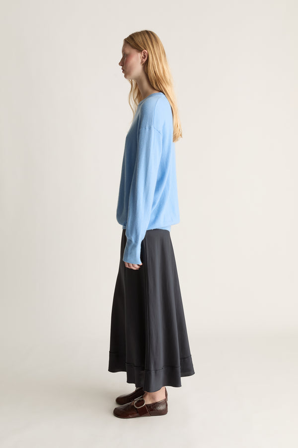 A woman stands in profile wearing a light blue long-sleeve knit top, a navy silk skirt, and brown leather loafers against a plain off-white studio background.