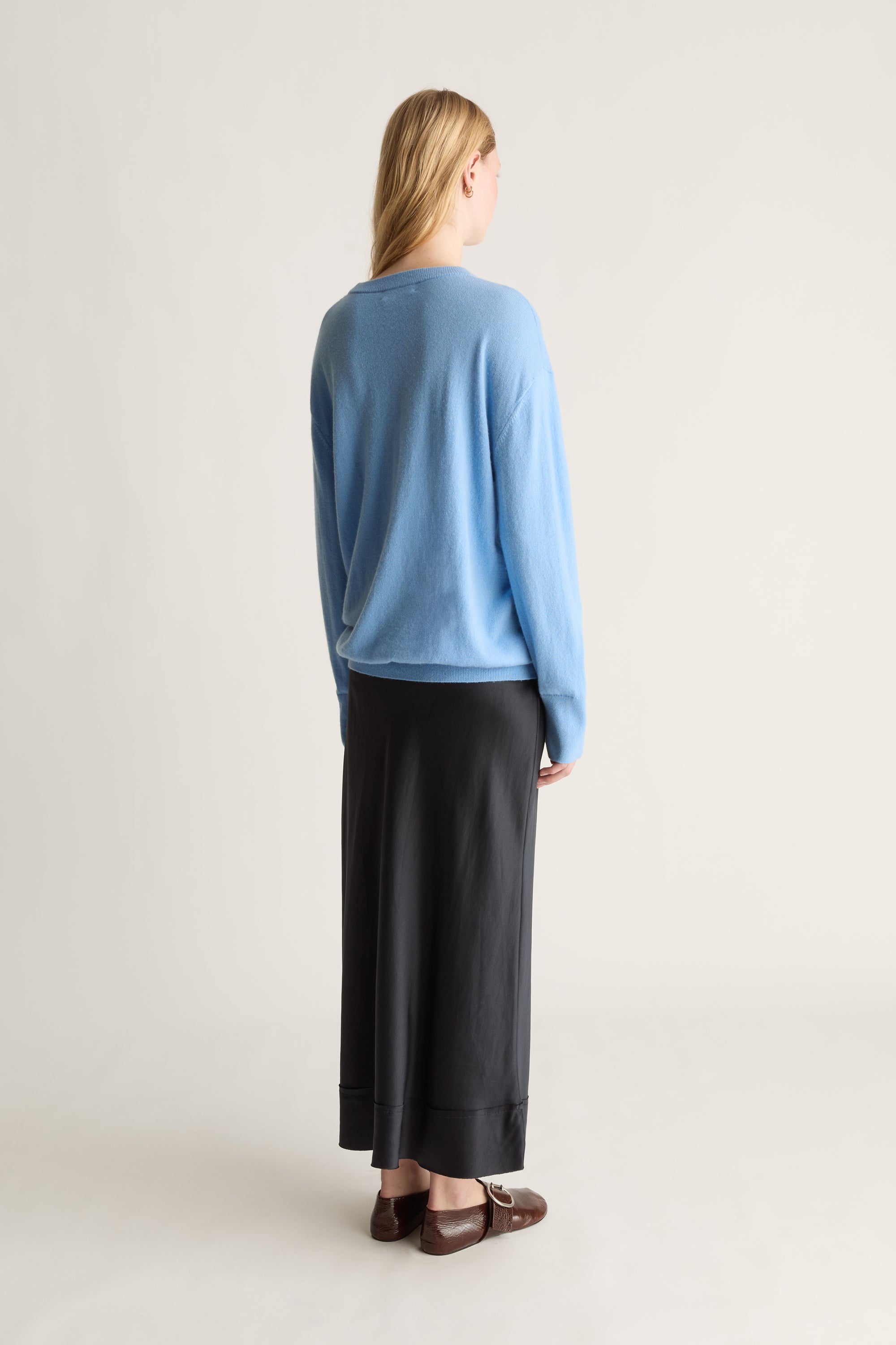 A woman stands with her back to the camera wearing a light blue long-sleeve knit sweater and a long black skirt, paired with brown leather loafers, against a plain off-white studio background.