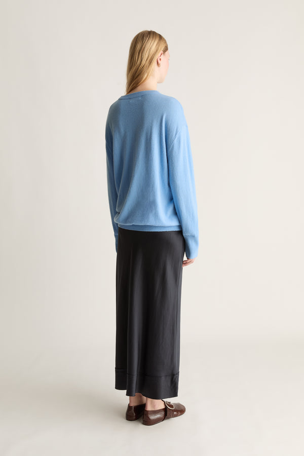 A woman stands with her back to the camera wearing a light blue long-sleeve knit sweater and a long black skirt, paired with brown leather loafers, against a plain off-white studio background.