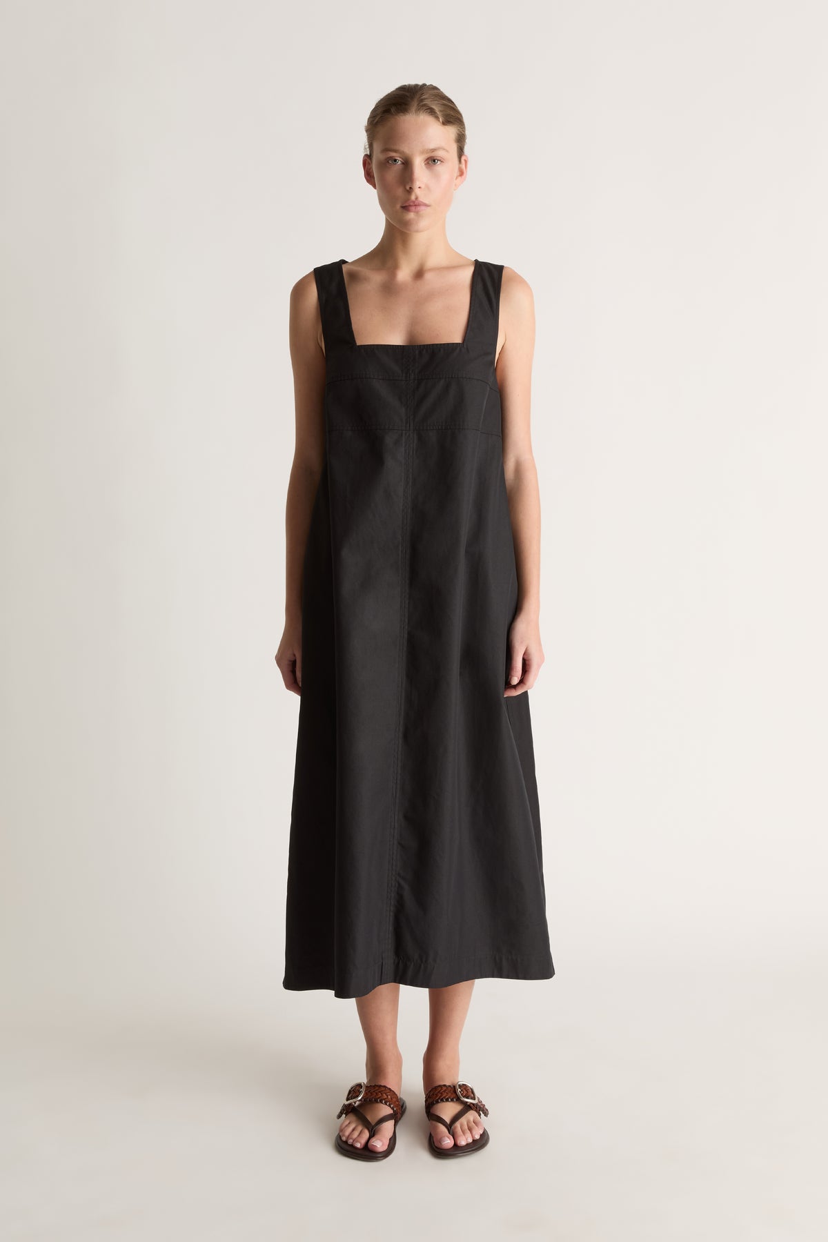 Front view of a black A-line midi dress with a square neckline, wide straps and topstitched yoke seams.