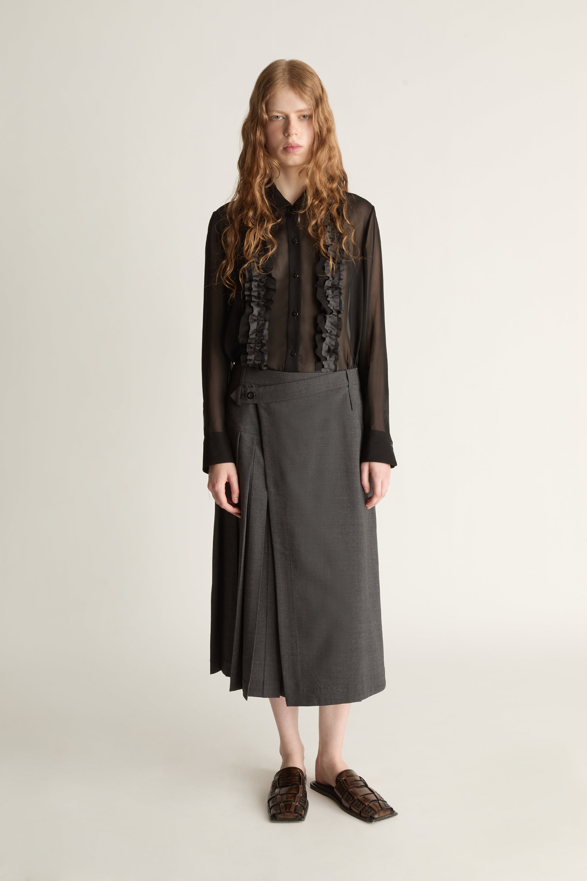 Person wearing a black blouse with lace details and a gray pleated skirt on a white background