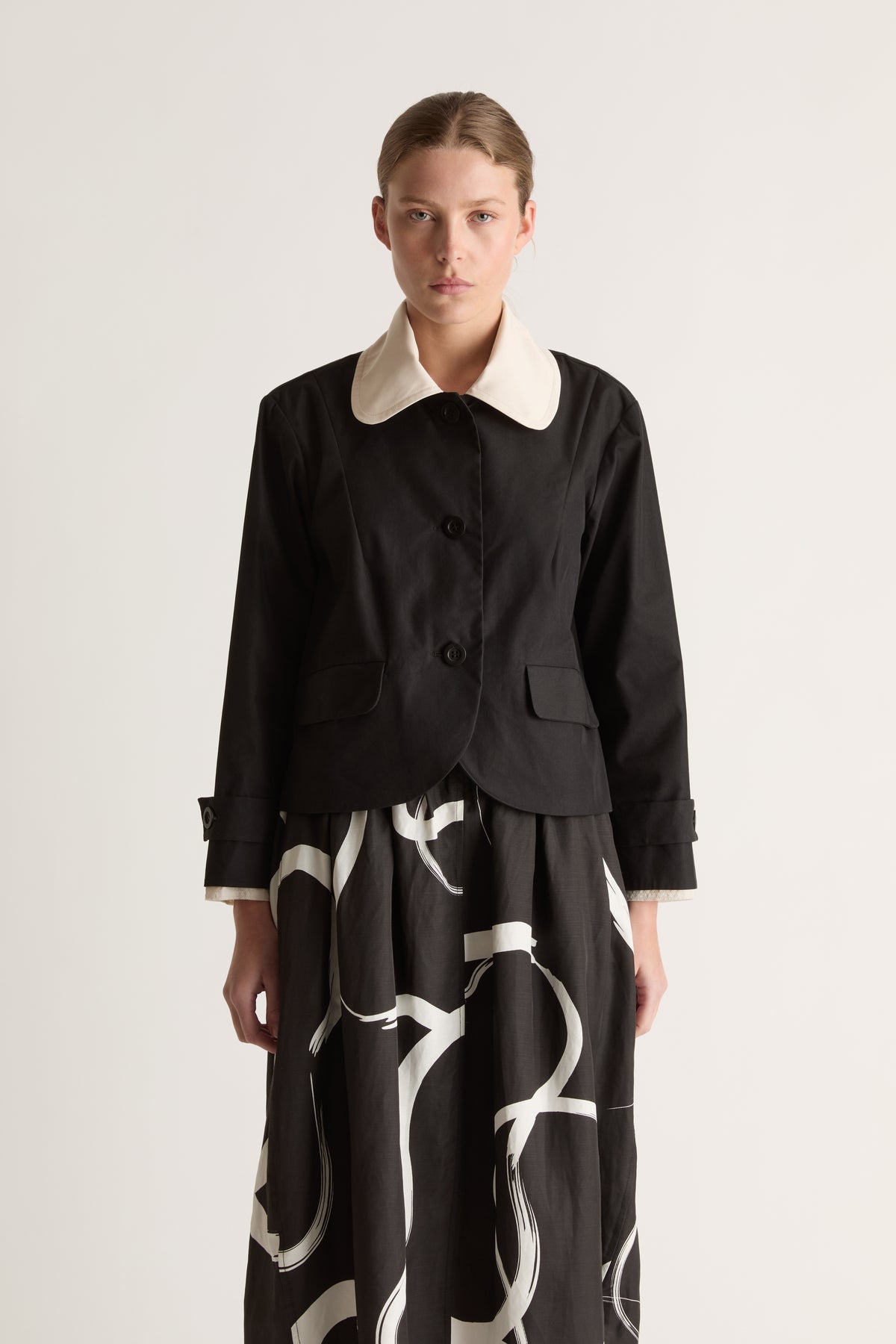 Close-up front view of a black jacket with a rounded cream collar, button front and flap pockets.