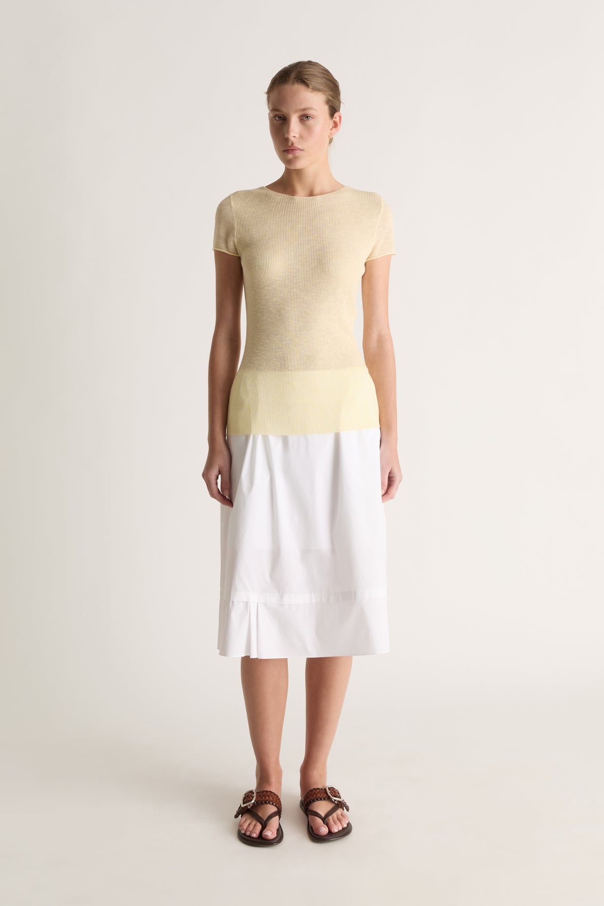 Front view of a woman wearing a pale yellow knit tee and white midi skirt against a white studio background