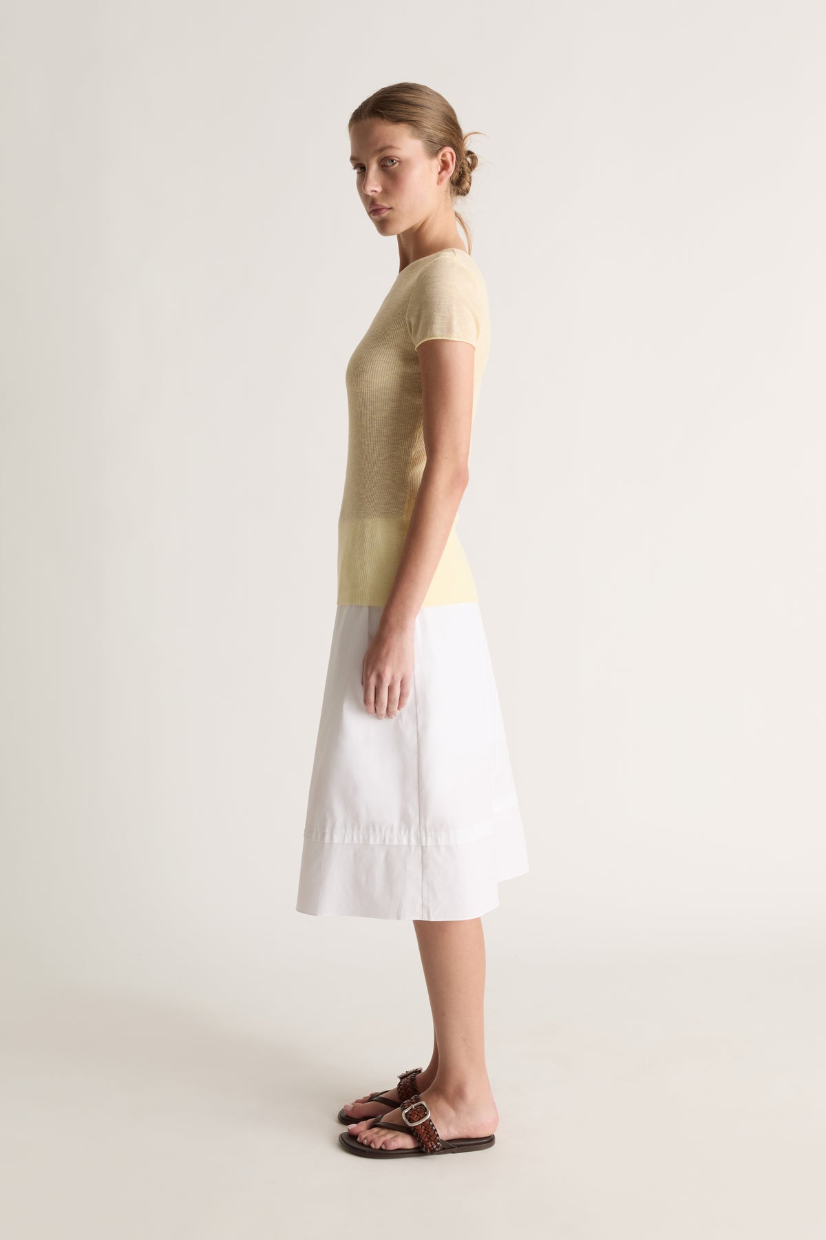 Side view of a woman with a bun in a pale yellow knit tee and white midi skirt with brown leather slides