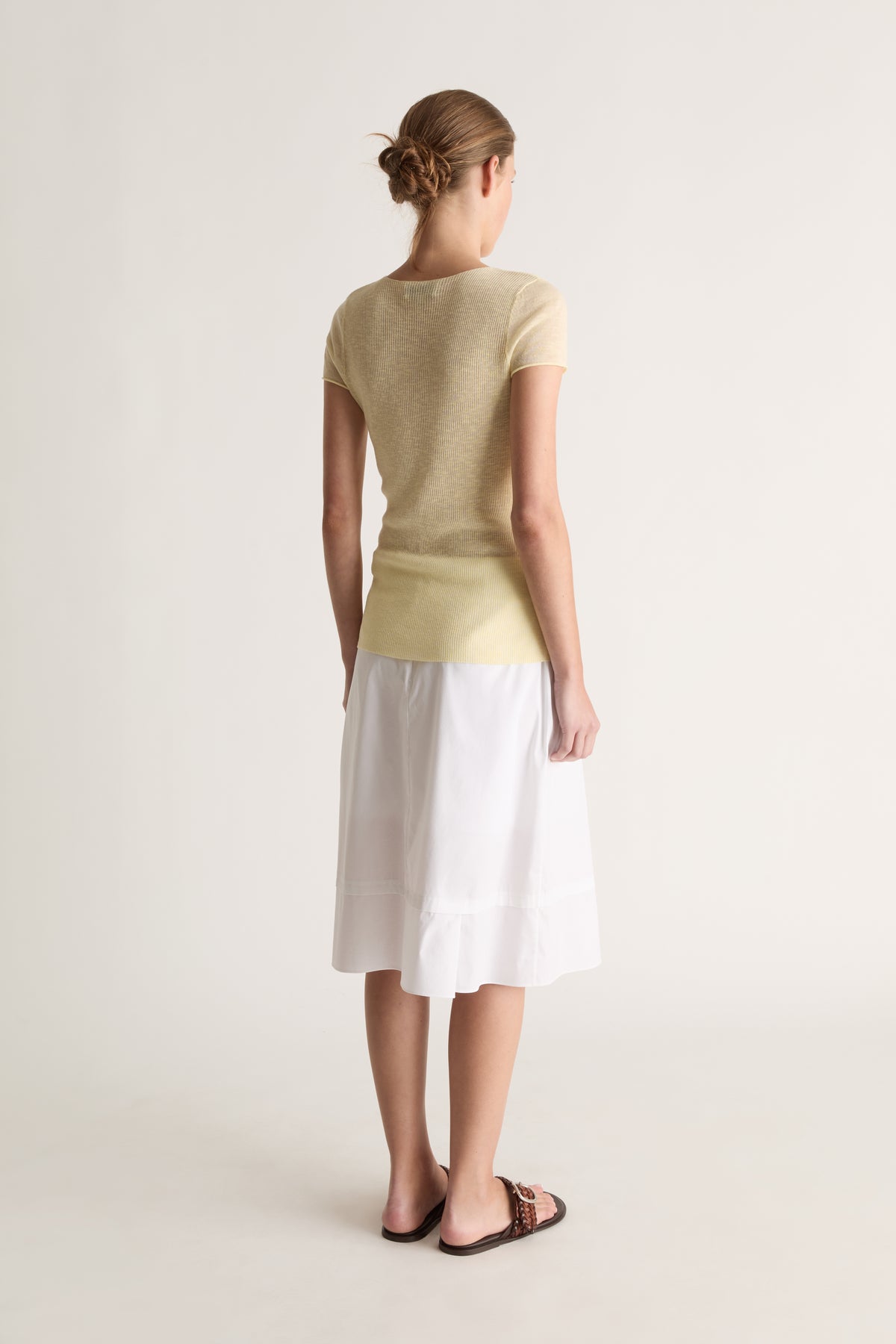 Back view of a woman with a low bun in a pale yellow short-sleeve knit tee and white skirt with brown slides
