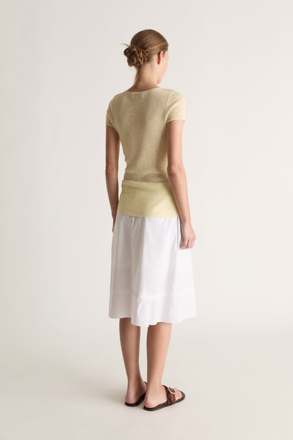 Back view of a woman with a low bun in a pale yellow short-sleeve knit tee and white skirt with brown slides