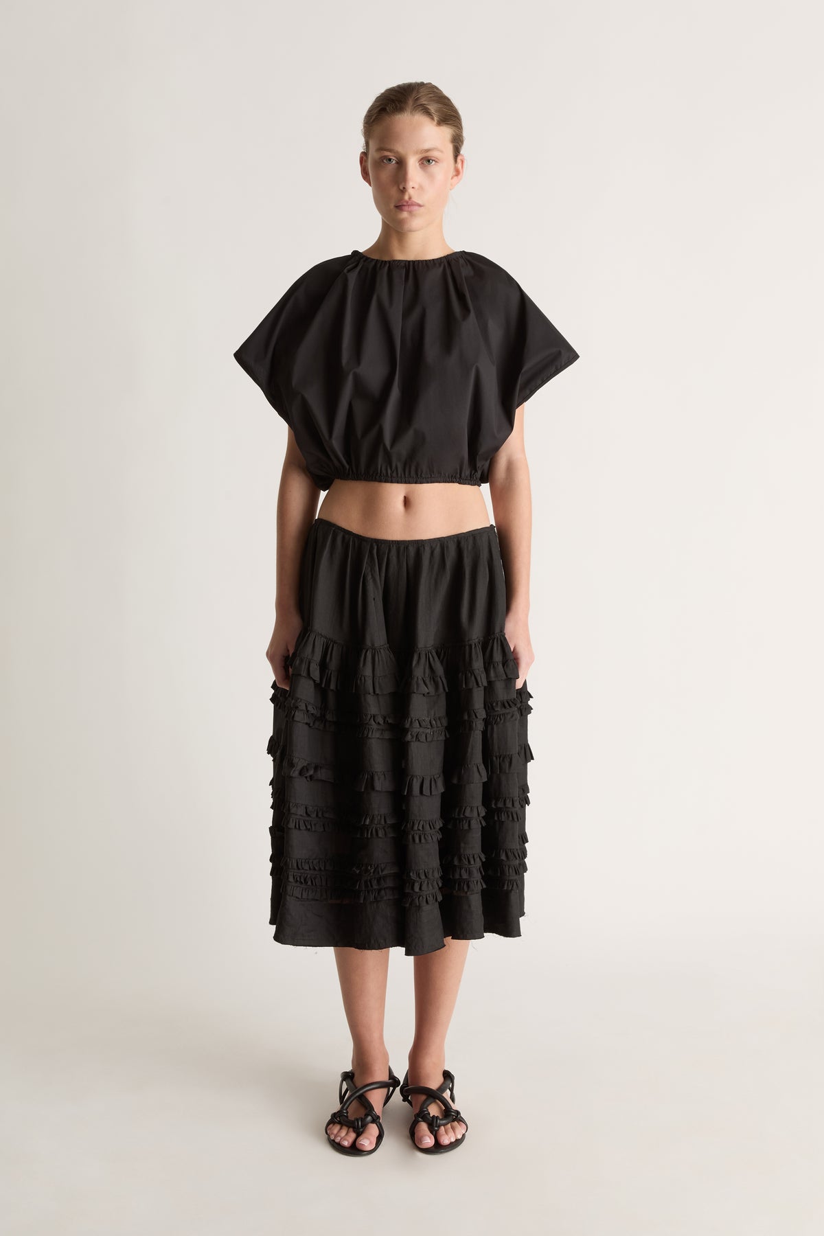 Front view of a woman wearing a black cropped top and tiered ruffle midi skirt against a white studio background