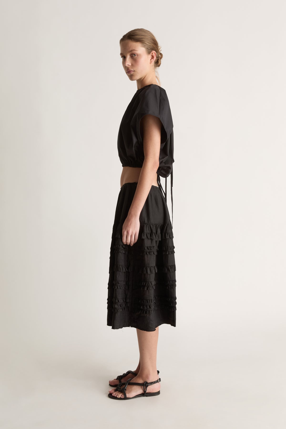 Side view of a woman with a low bun in a black cropped top with tie detail and a black tiered ruffle skirt