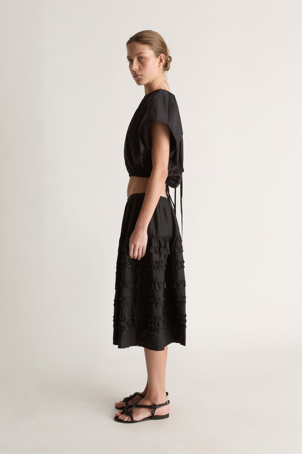 Side view of a woman with a low bun in a black cropped top with tie detail and a black tiered ruffle skirt