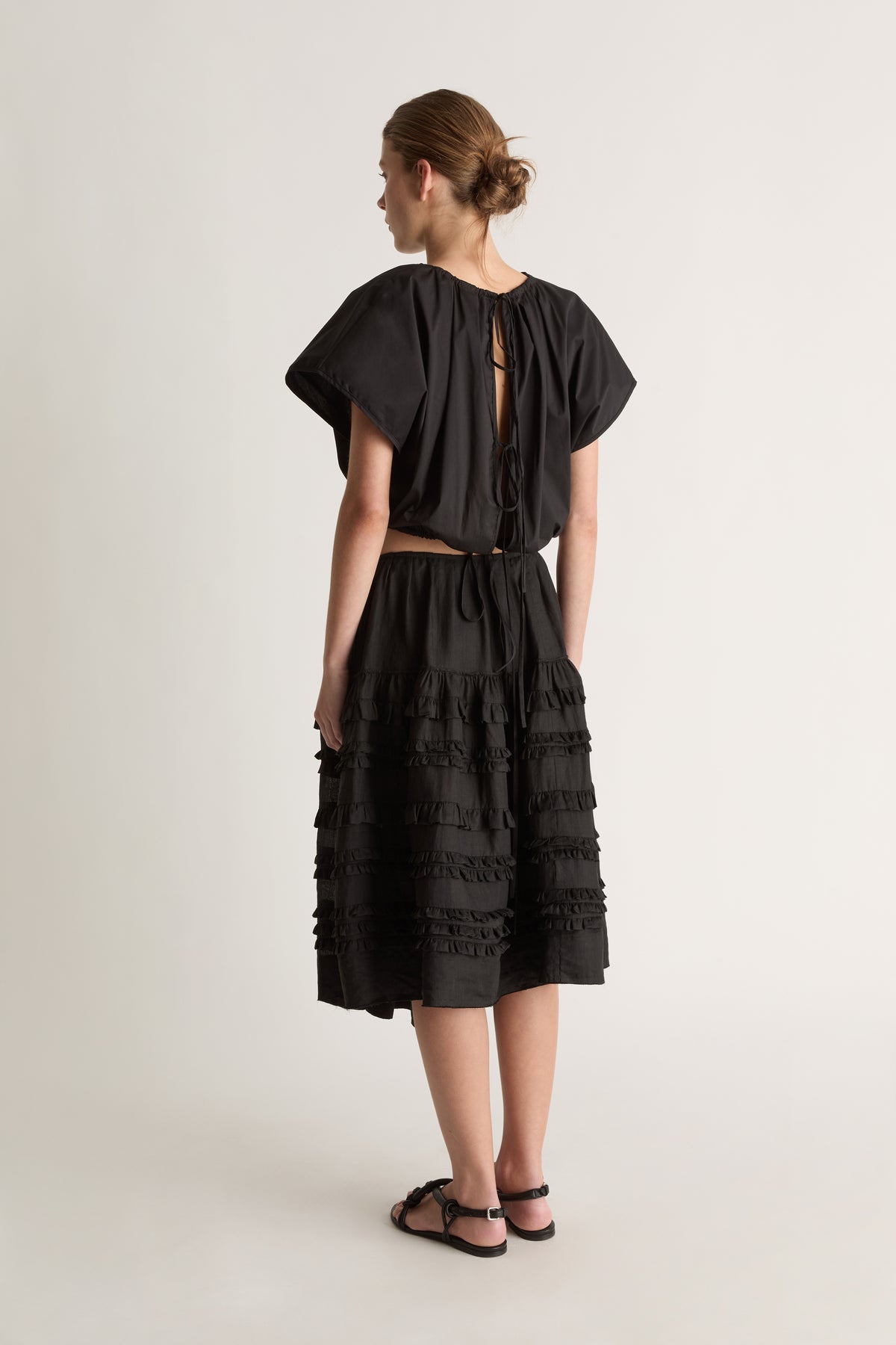 Back view of a woman with a bun in a black cropped top with keyhole tie back and a black ruffle-tiered midi skirt
