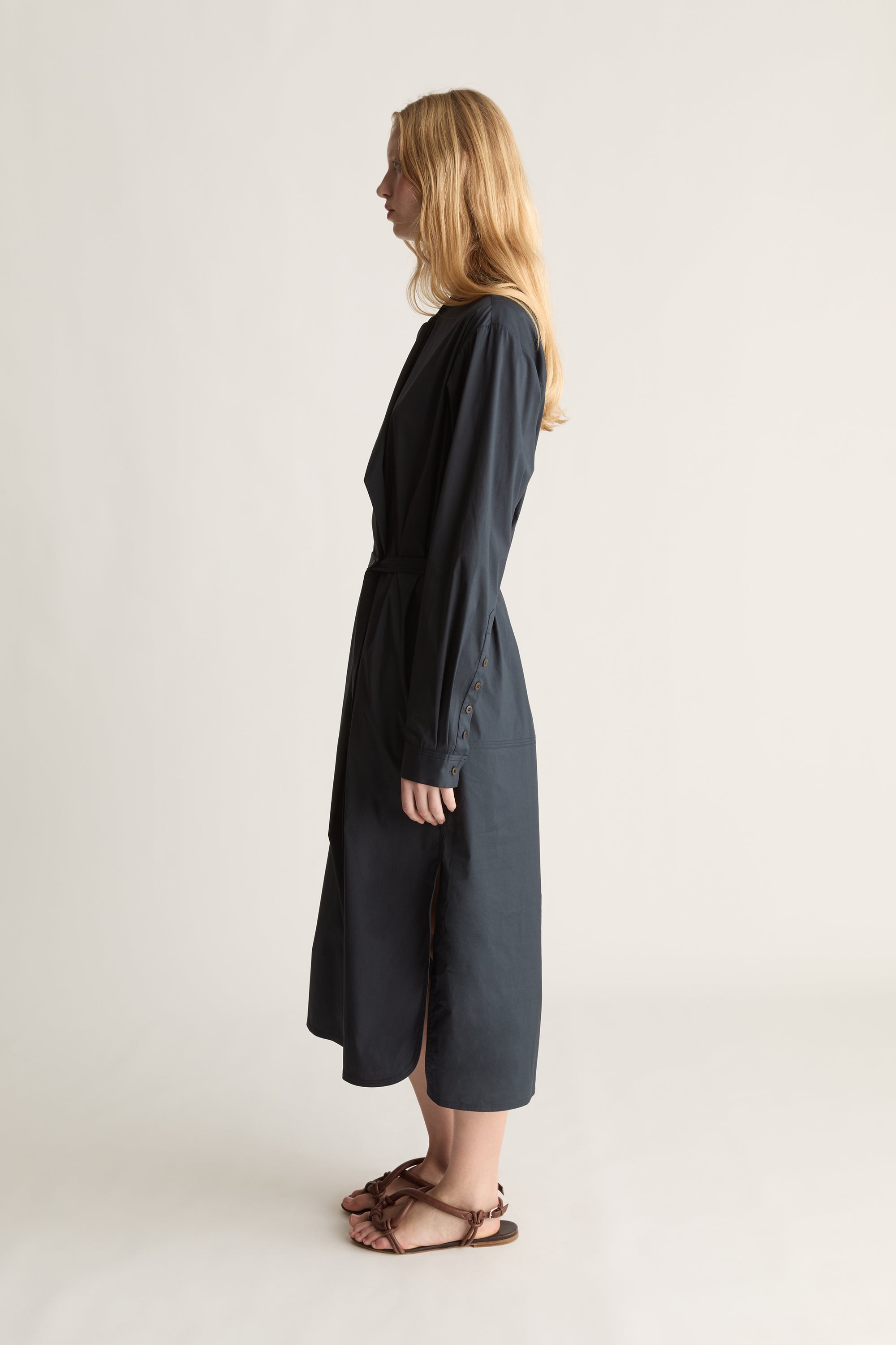 A woman stands in profile wearing a navy blue midi dress with long sleeves and brown strappy flat sandals against a plain off-white studio background.