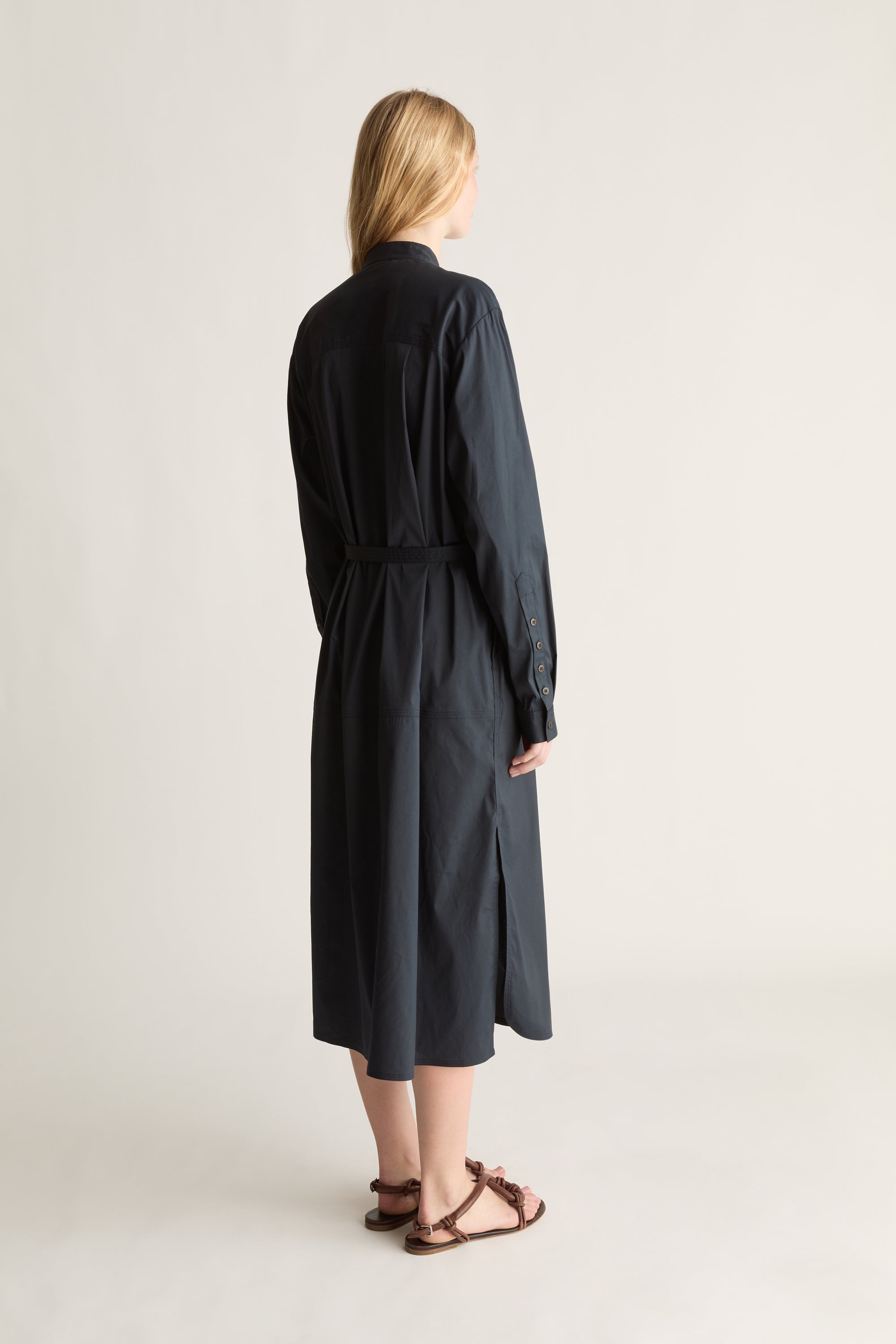 A woman shown from the back wearing a long dark navy midi shirt dress with a tie waist belt, paired with brown strappy flat sandals, against a plain light gray studio background.