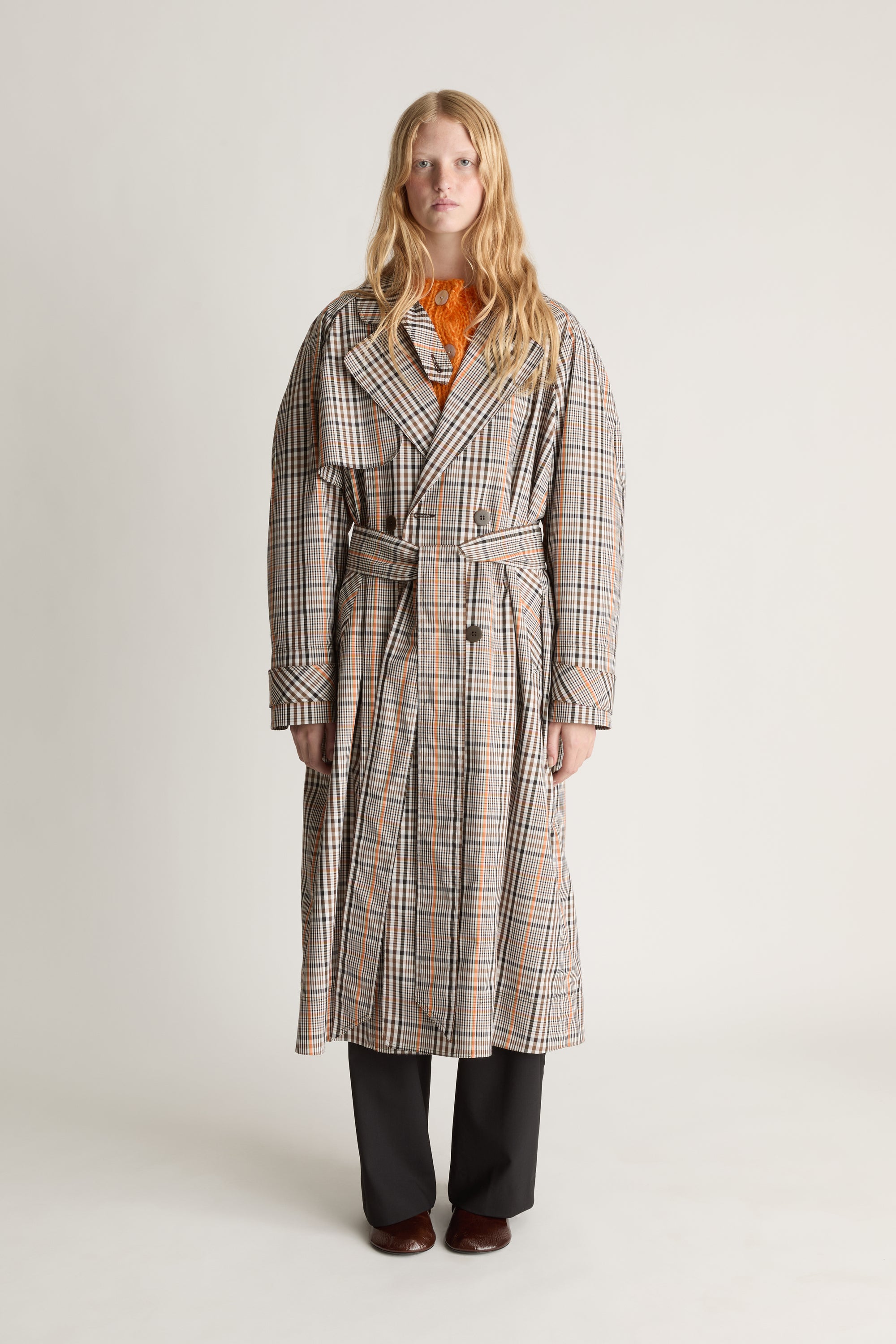 A woman stands against a plain light gray studio background wearing a long beige plaid coat with a belt, an orange top underneath, dark wide-leg pants, and brown shoes.