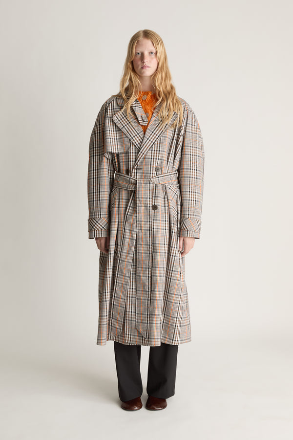 A woman stands against a plain light gray studio background wearing a long beige plaid coat with a belt, an orange top underneath, dark wide-leg pants, and brown shoes.