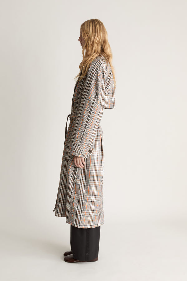 A woman stands in profile wearing a long plaid wrap coat in beige, gray, and brown tones, paired with dark wide-leg pants, against a plain light studio background.