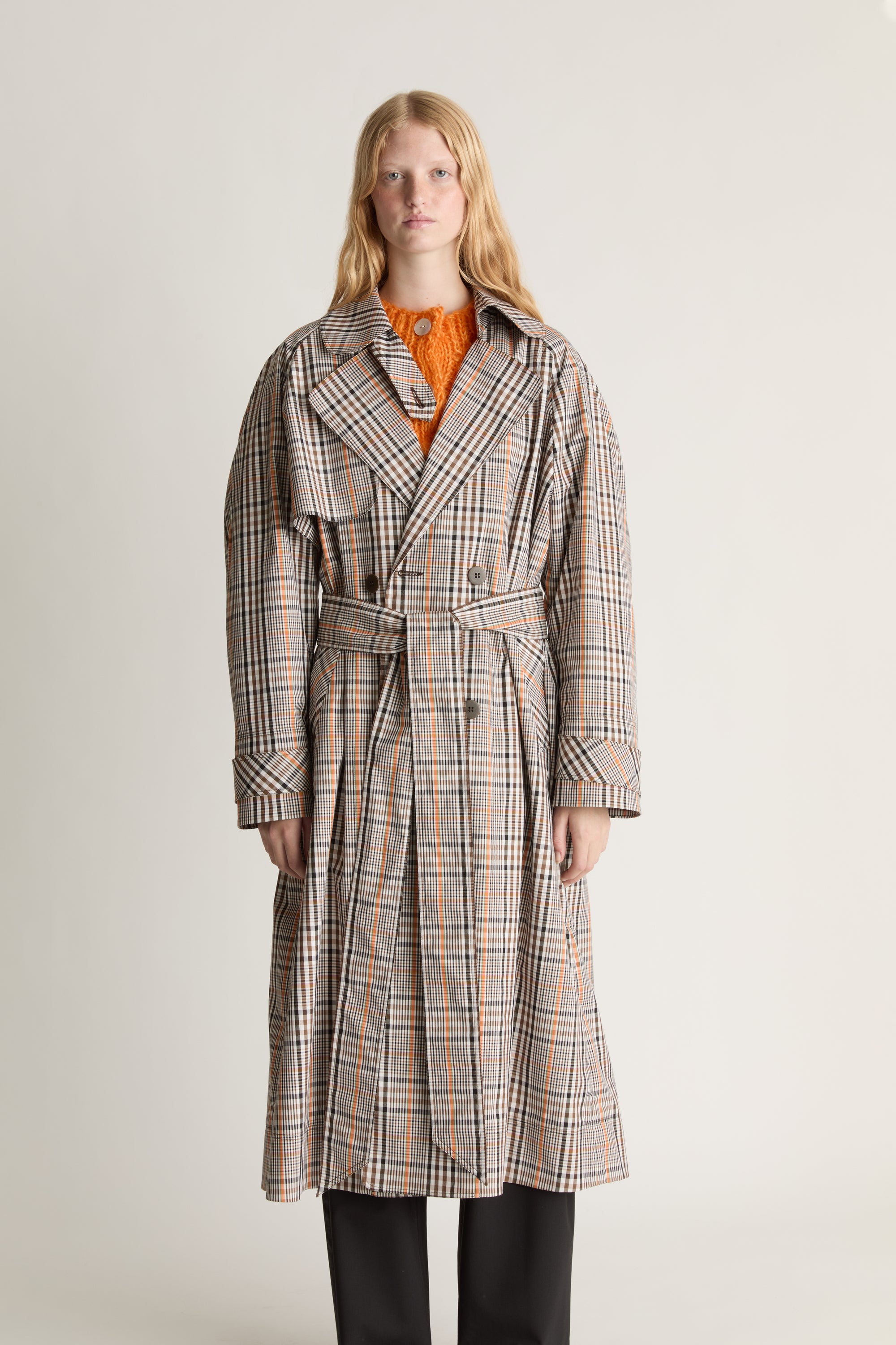 Woman facing the camera wearing a plaid coat with orange accents and black torusers  on a plain background