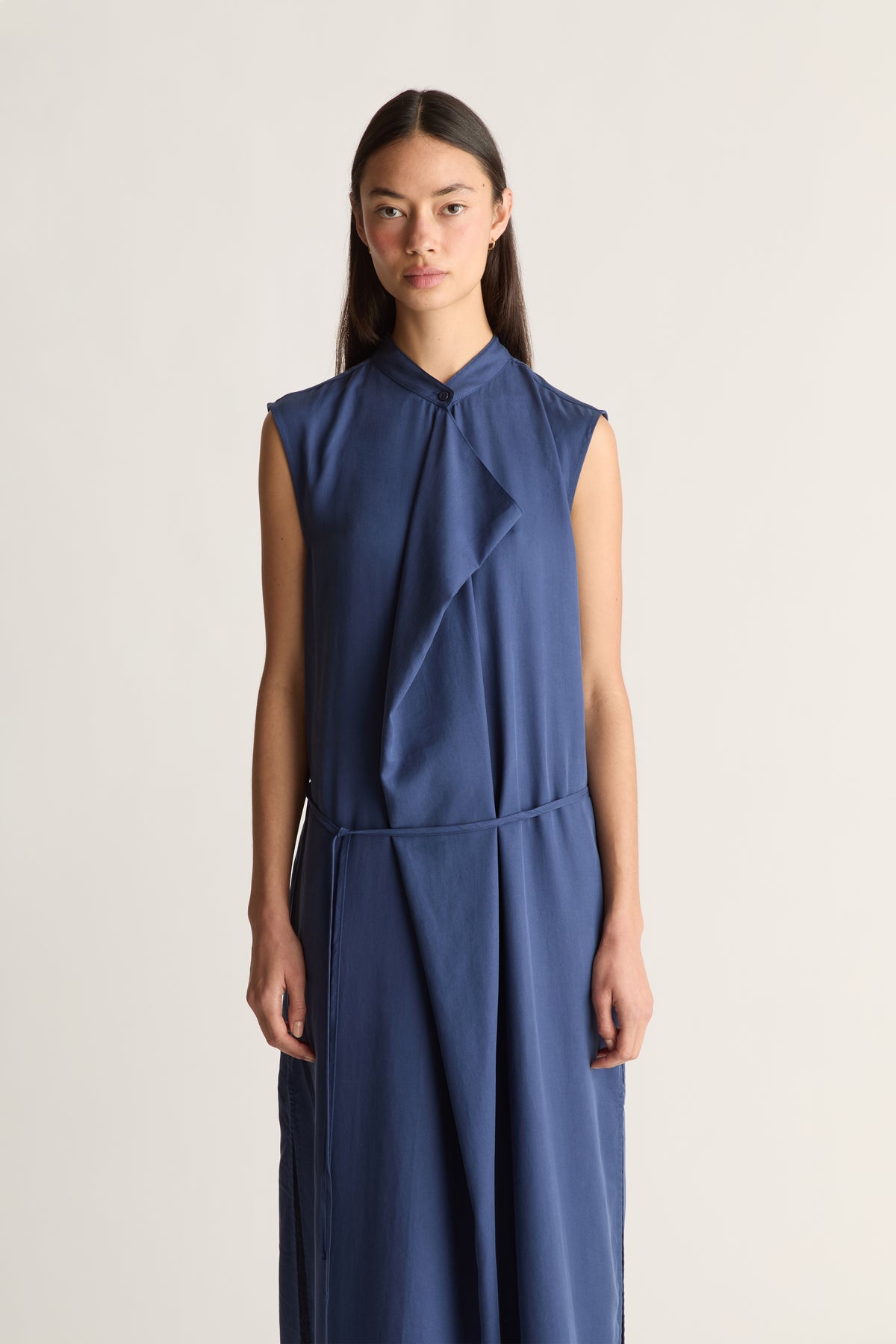 Close up of a woman wearing a sleeveless, high-neck blue dress with a draped wrap-style front and a matching tie belt, set against a plain light background.