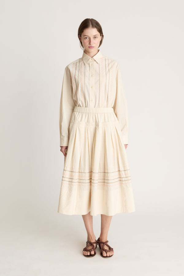 A woman wearing a beige shirt with a beige skirt that has embroidered details.