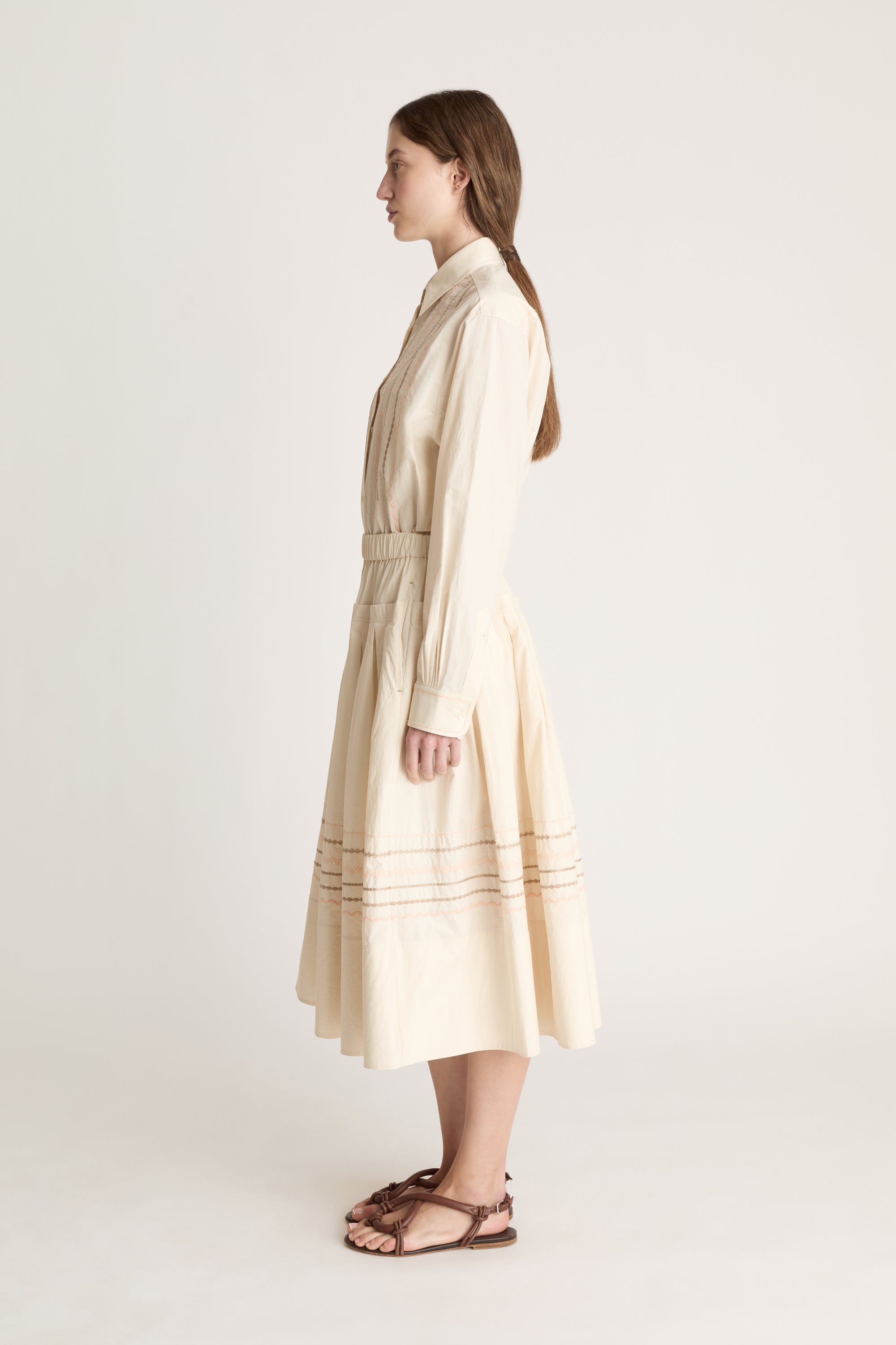A woman wearing a beige shirt with a beige skirt that has embroidered details on a white background