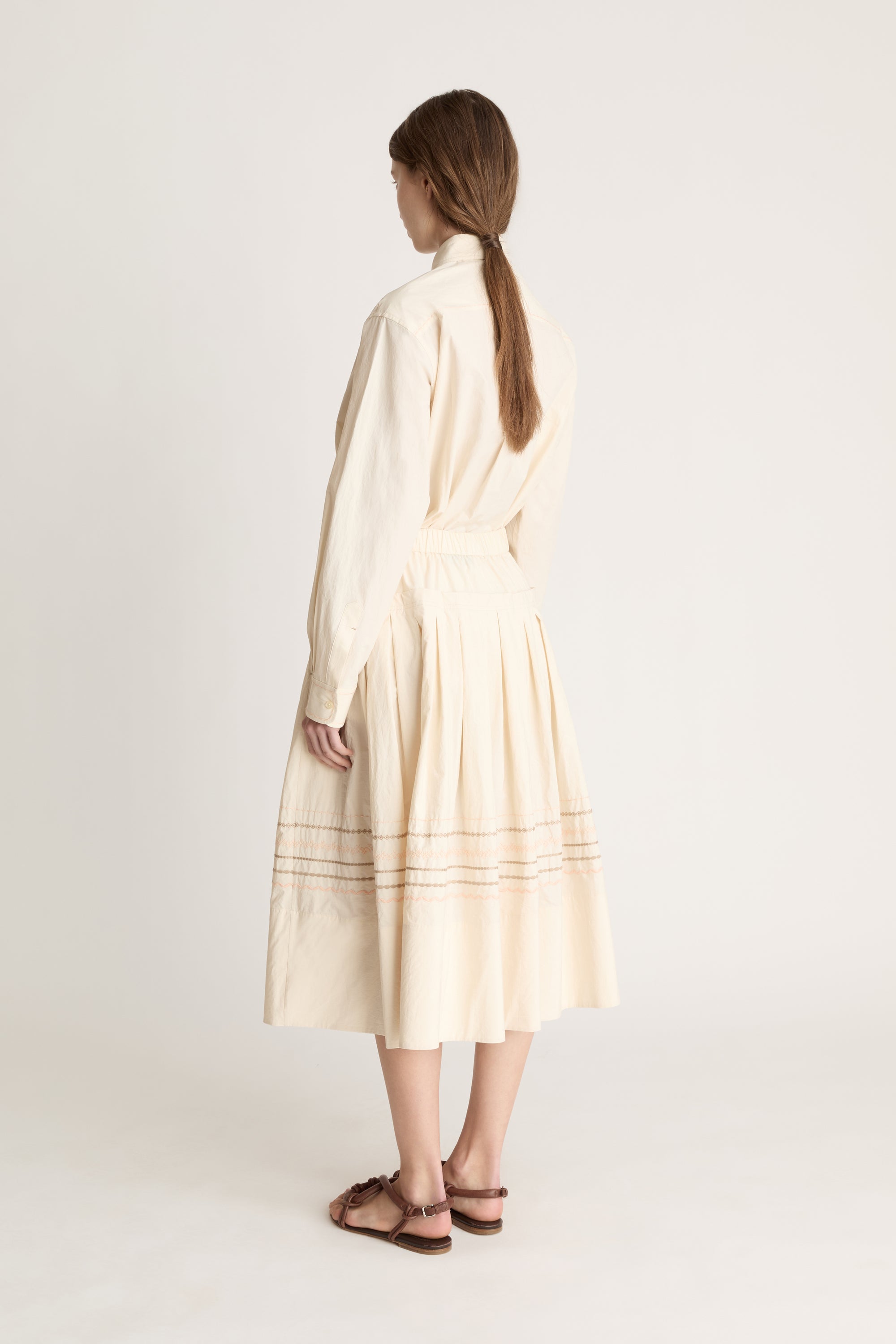 A woman wearing a beige shirt with a beige skirt that has embroidered details on a white background