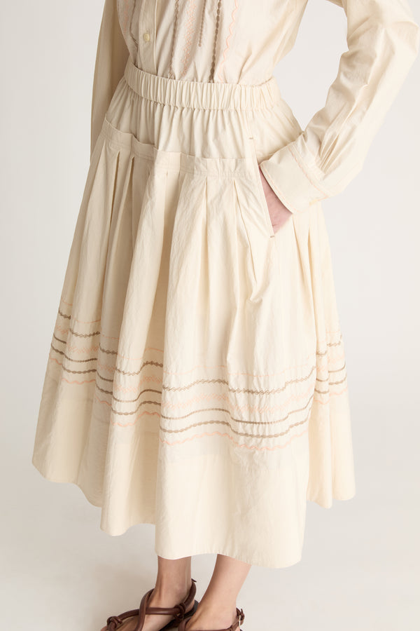 A woman wearing a beige shirt with a beige skirt that has embroidered details on a white background