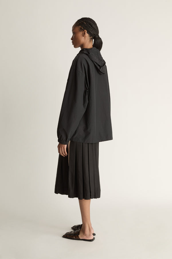 Woman wearing a black jacket with a white zipper and a black pleated skirt on a light background