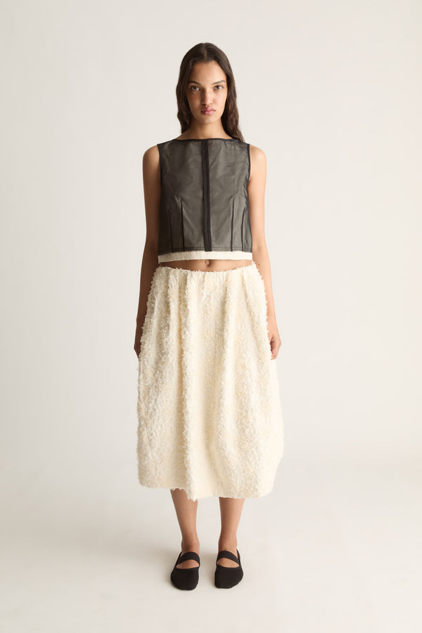 Front view of a cropped sleeveless sheer black top with vertical seams and a cream hem, worn with a cream skirt.