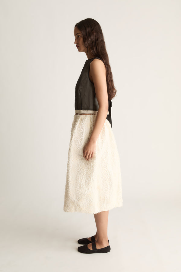 Side view of a cropped sleeveless sheer black top with back ties, worn with a textured cream skirt.