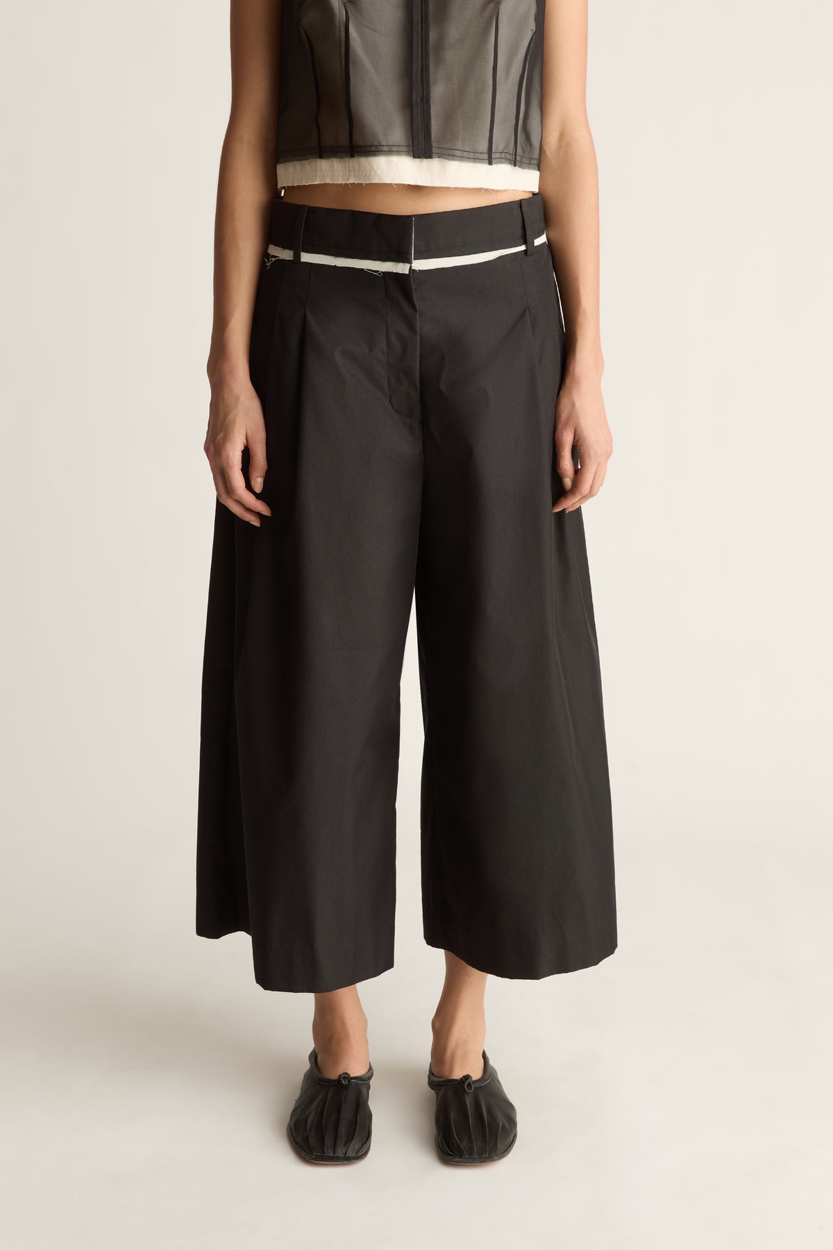 Close-up front view of black wide-leg cropped pants with front pleats and a cream-edged waistband.