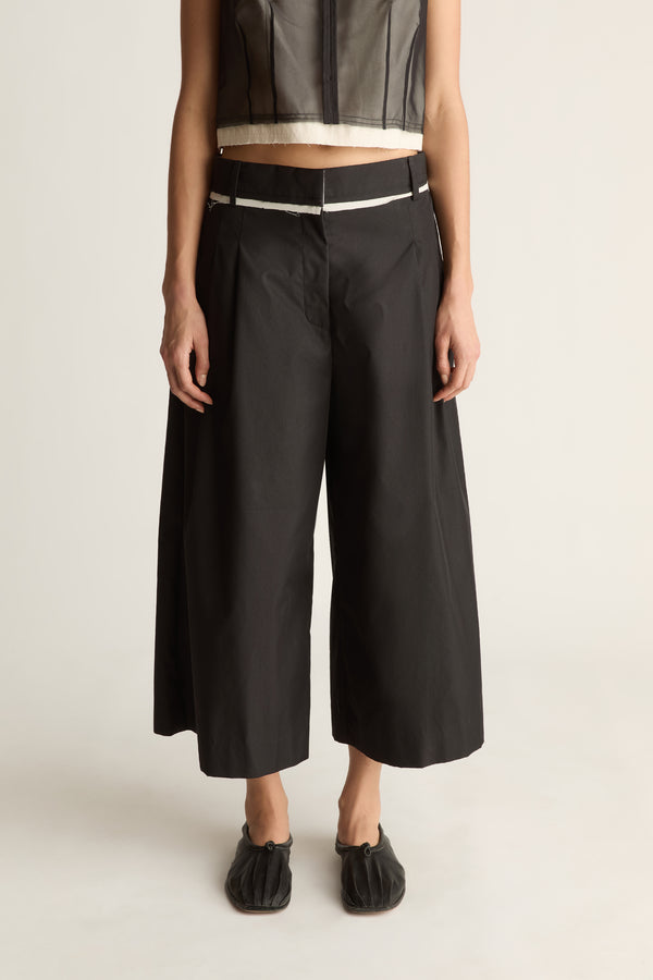 Close-up front view of black wide-leg cropped pants with front pleats and a cream-edged waistband.