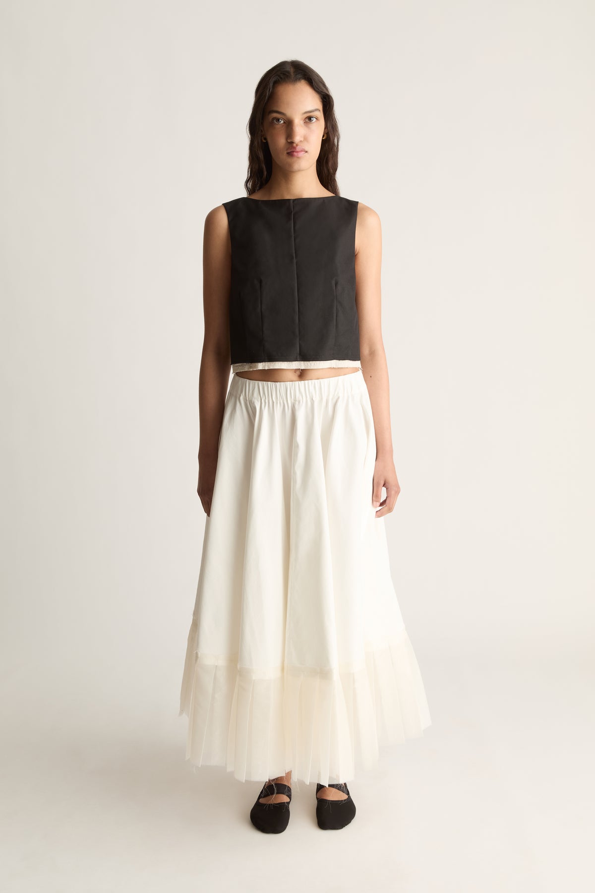 Front view of a cropped sleeveless black top with a high round neckline and seamed front, worn with a cream skirt.