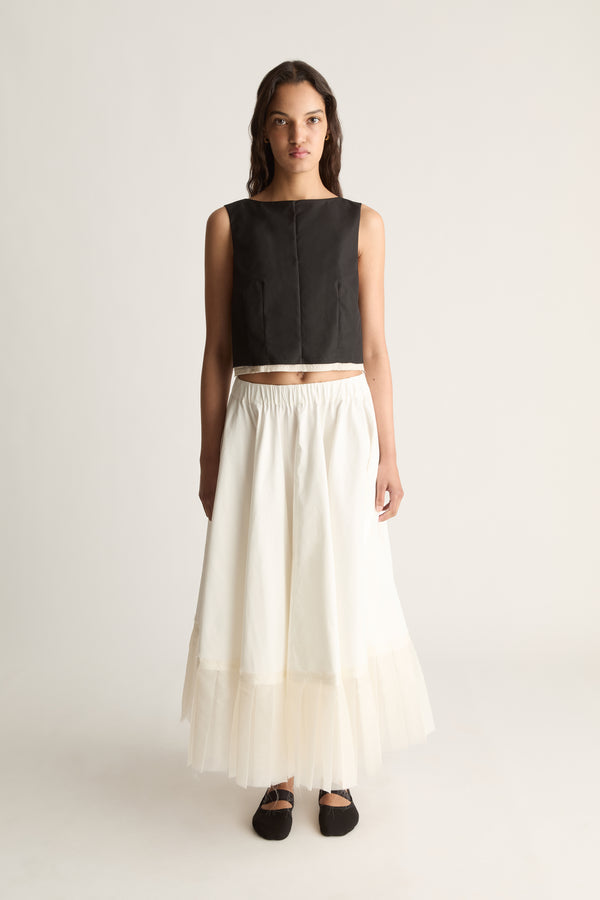 Front view of a cropped sleeveless black top with a high round neckline and seamed front, worn with a cream skirt.