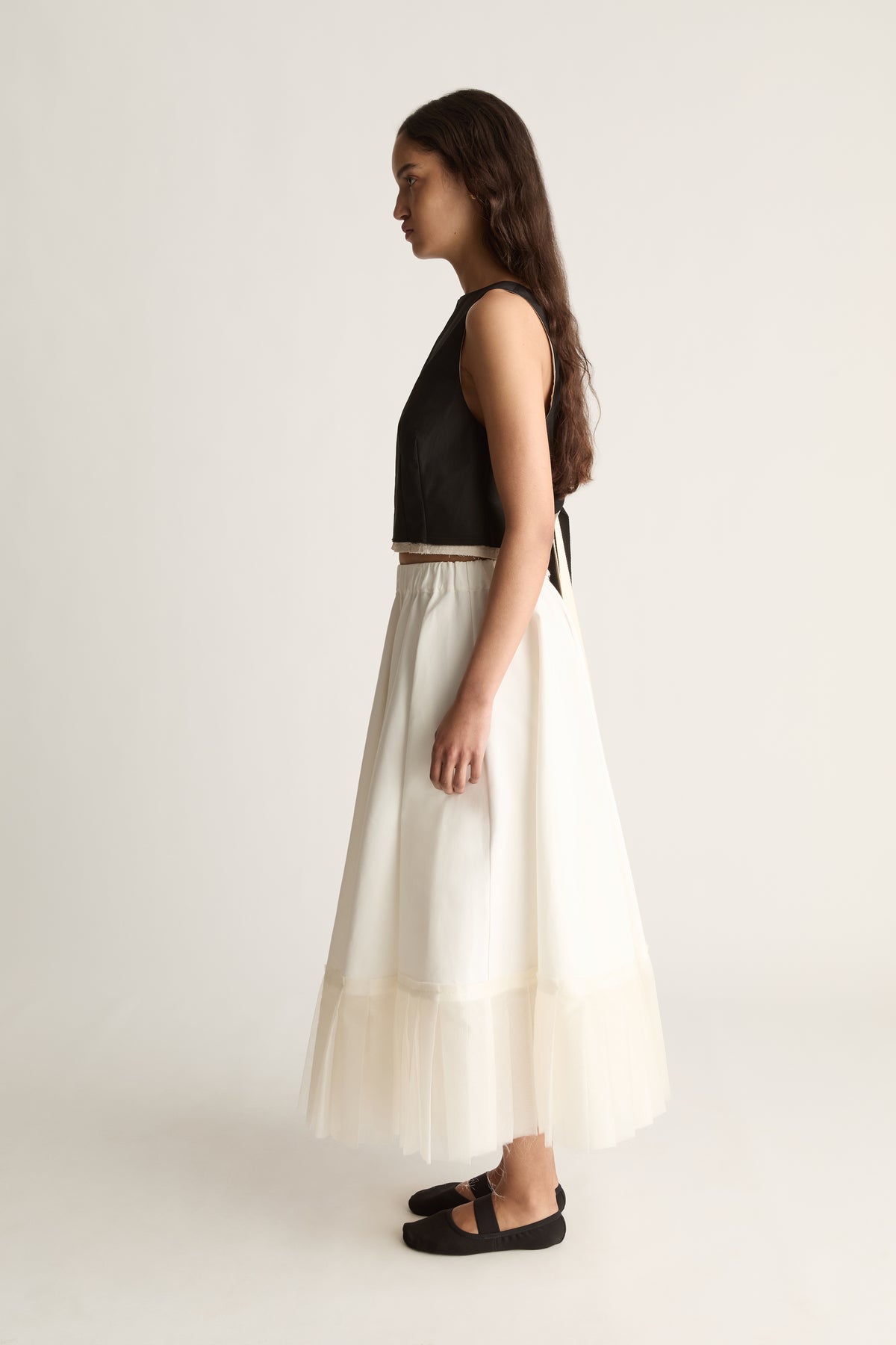 Side view of a cropped sleeveless black top with ties at the back, worn with a full cream skirt.