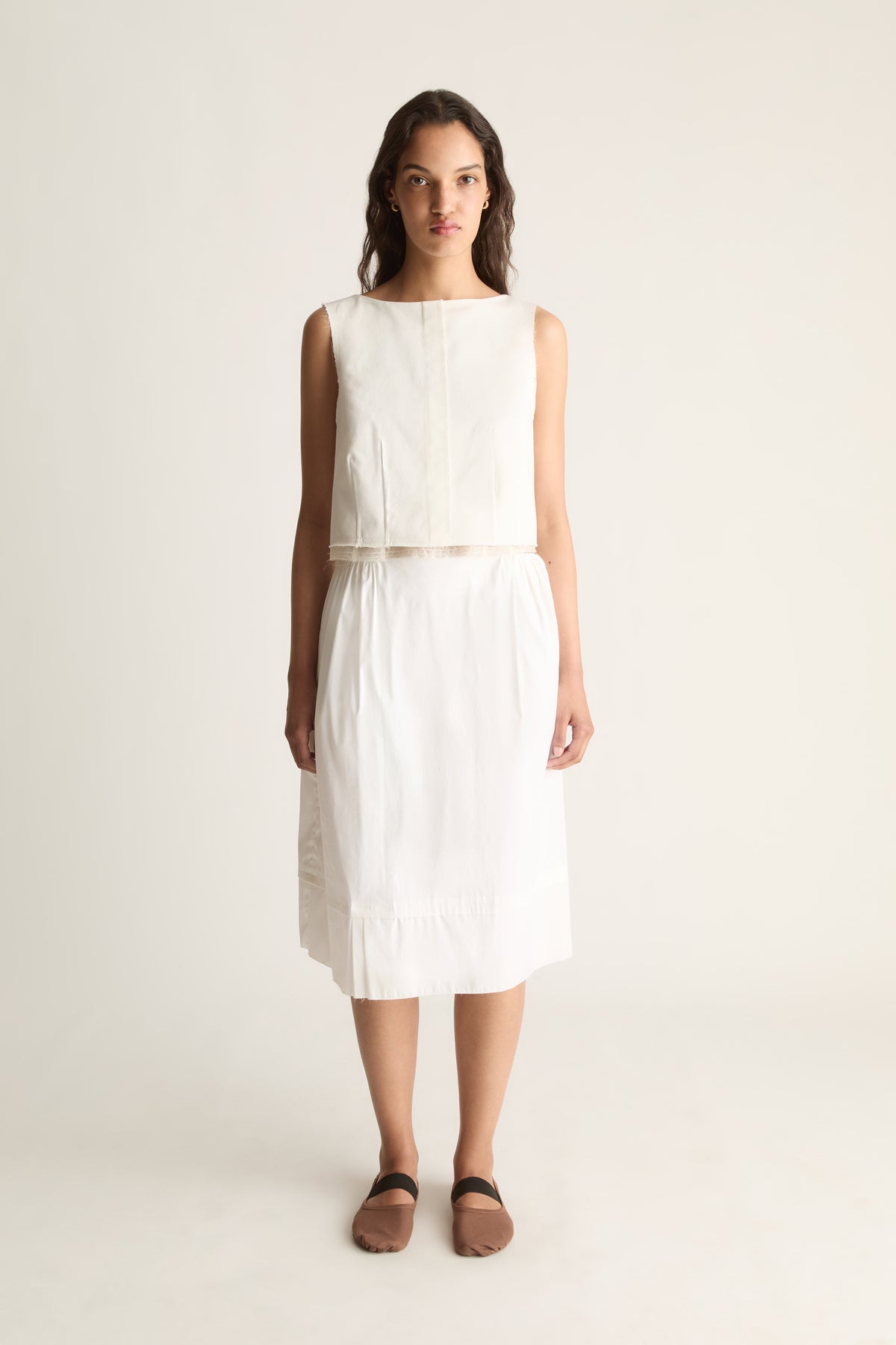 Front view of a cropped sleeveless cream top with a round neckline and seamed front, worn with a cream skirt.