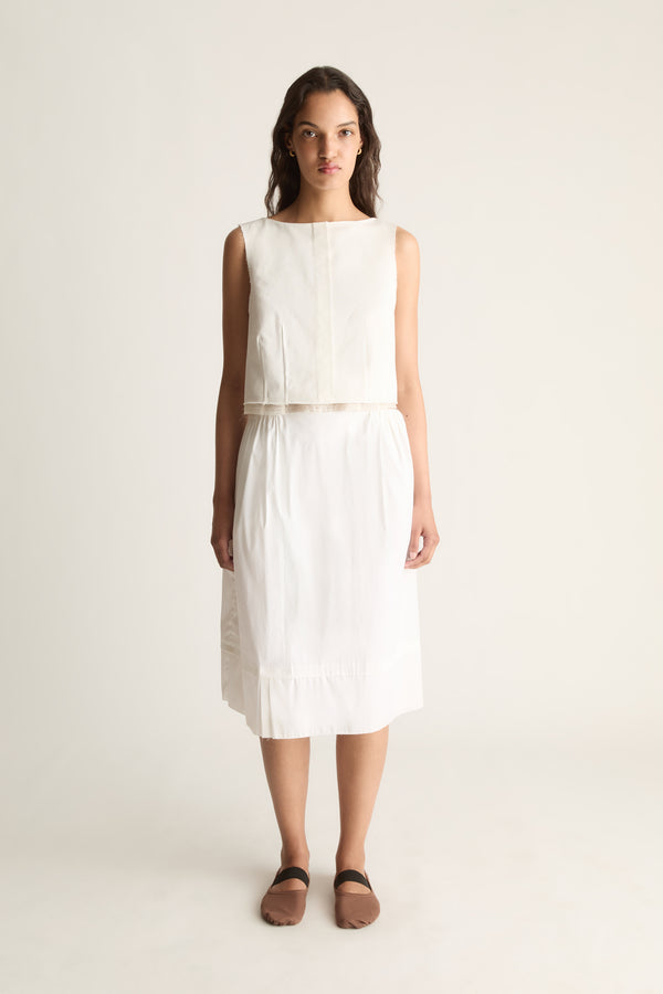 Front view of a cropped sleeveless cream top with a round neckline and seamed front, worn with a cream skirt.