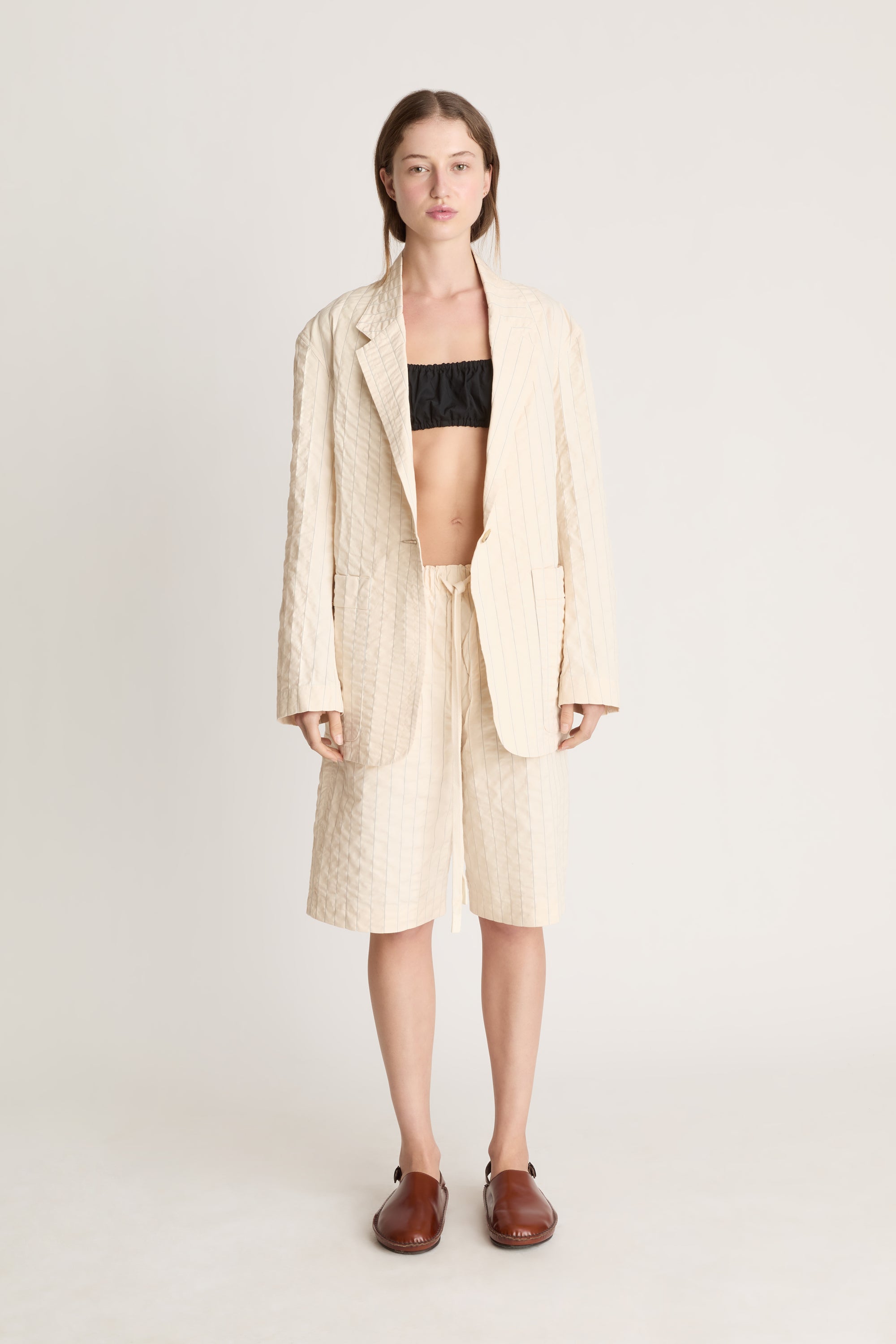 Woman wearing a cream blazer and shorts with a black bralette underneath on a white background.