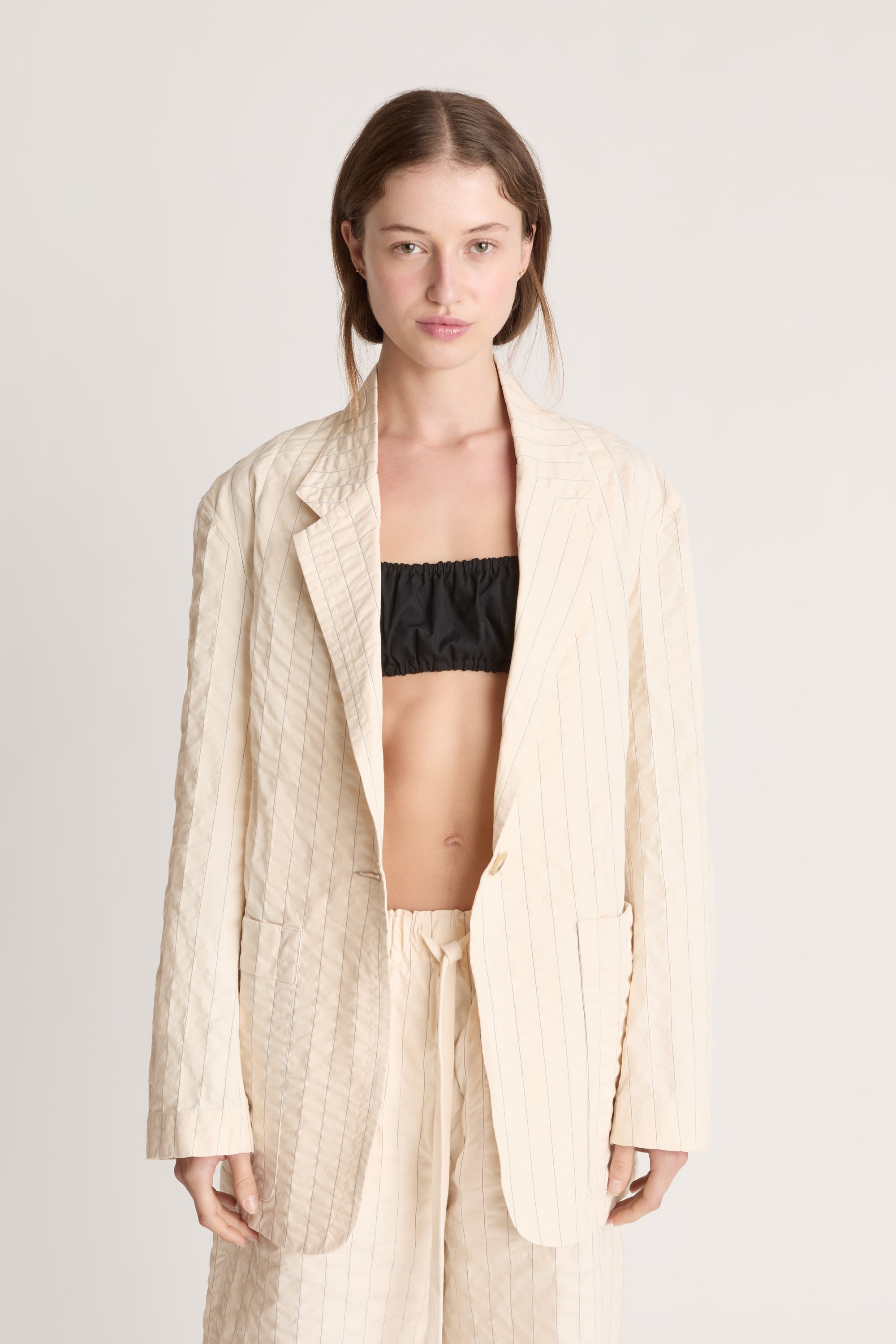Woman wearing a cream blazer and shorts with a black bralette underneath on a white background.
