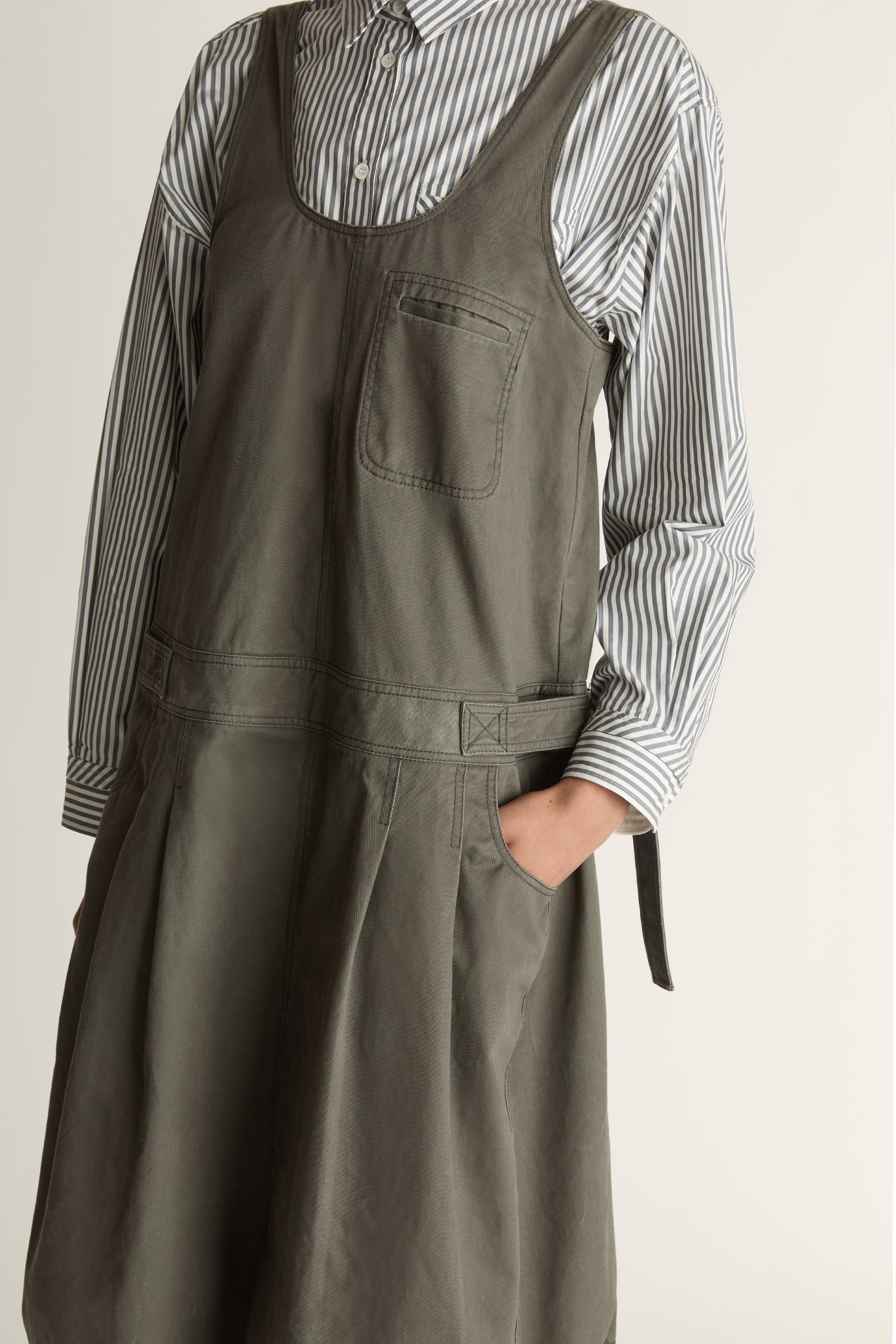 Woman wearing a green apron dress over a striped shirt on a plain background
