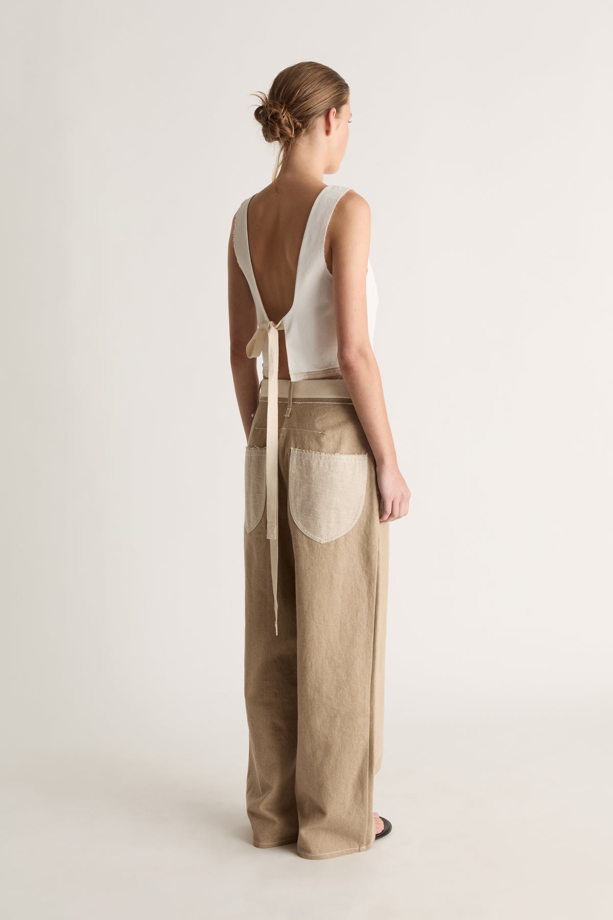 Back view of a woman with a bun in a white open-back top with long ties and beige jeans with cream back pockets