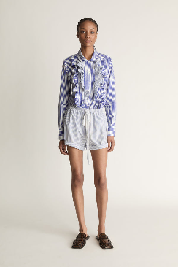 Person wearing a blue and white striped shirt with ruffled details and white shorts on a plain background