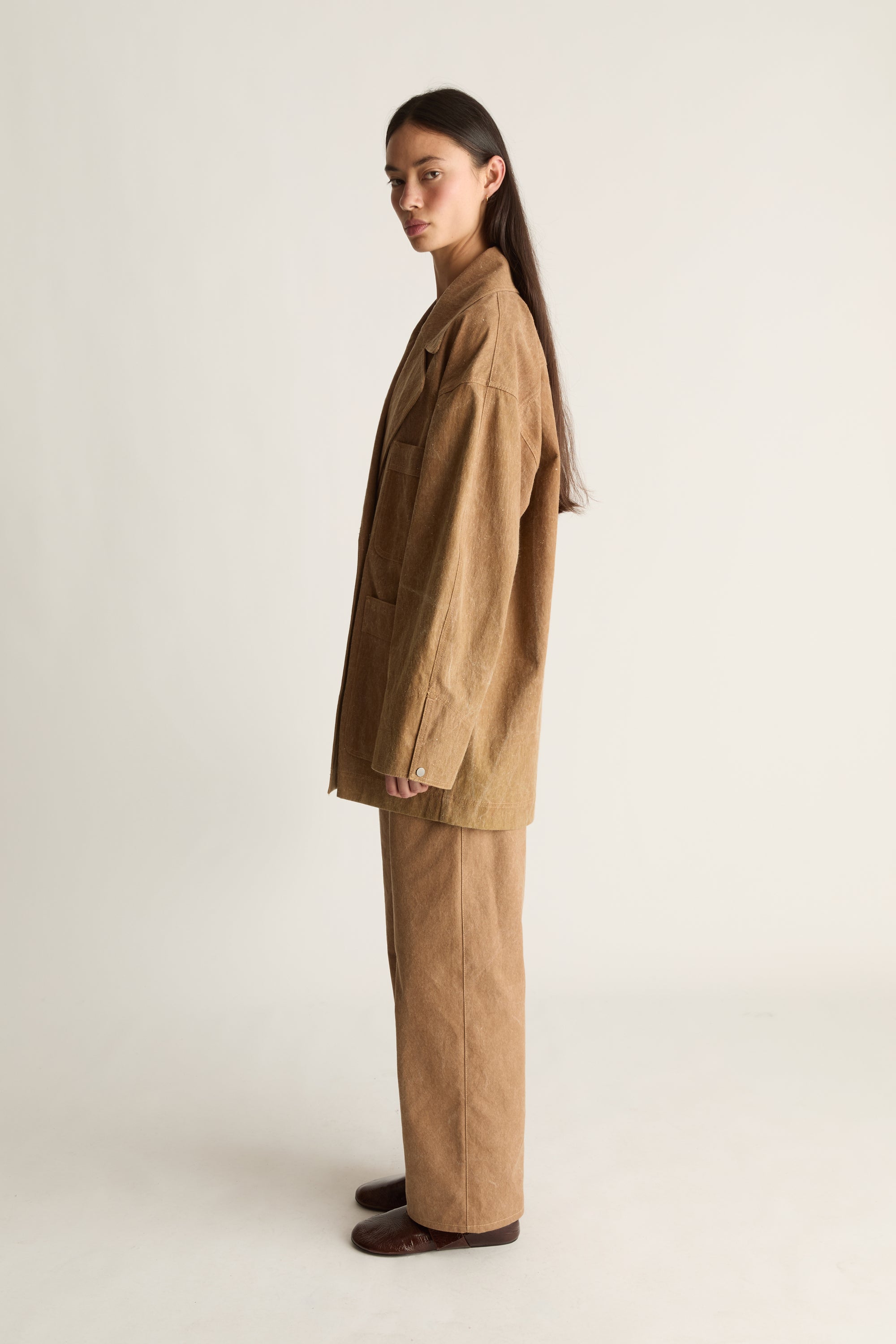 Woman wearing a brown coat and pants on a white background, facing the side but looking toward the front.