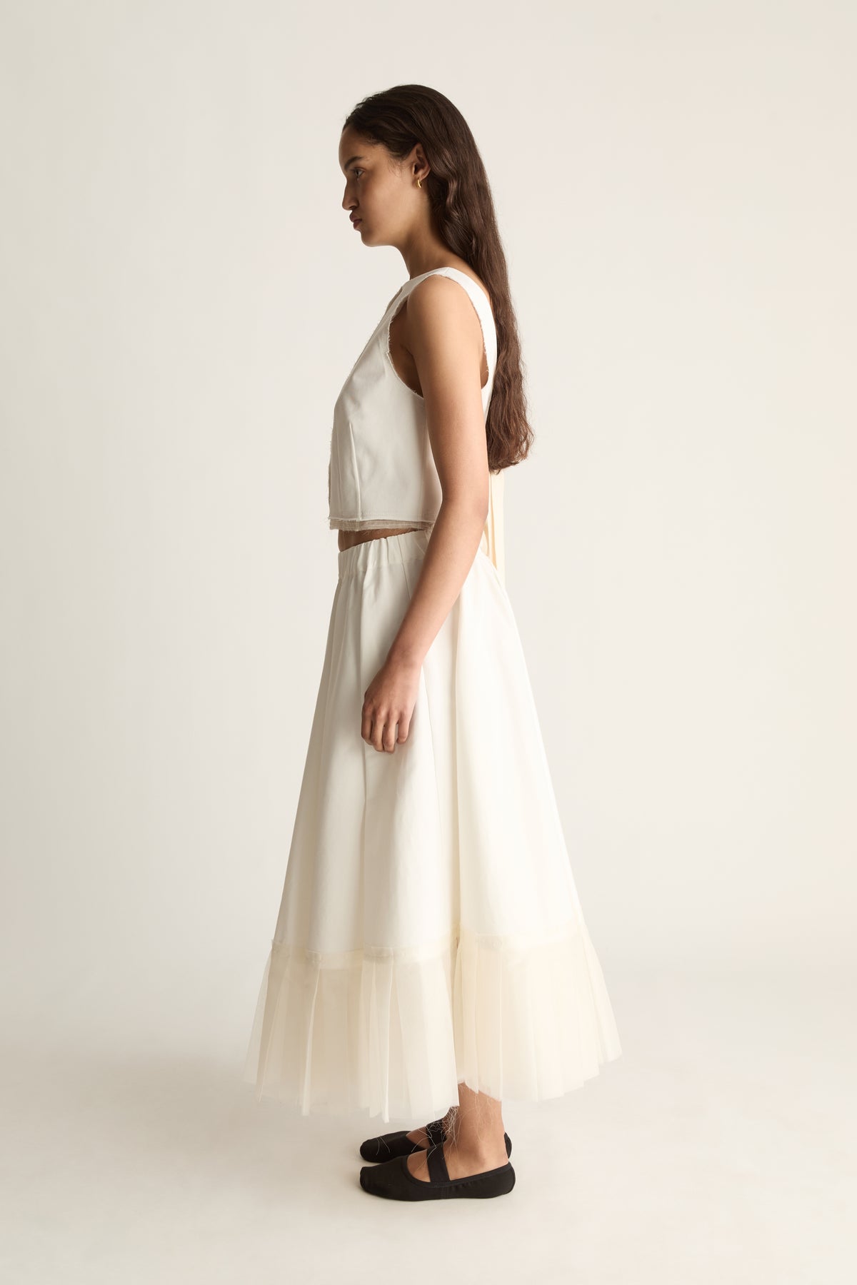 Side view of a full cream maxi skirt flaring out to a sheer layered hem, worn with a cream cropped top.