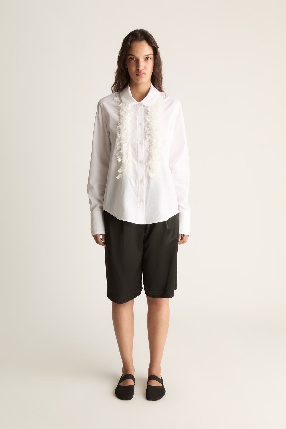 Front view of a woman wearing a white long-sleeve shirt with ruffle trim and black knee-length shorts against a white studio background