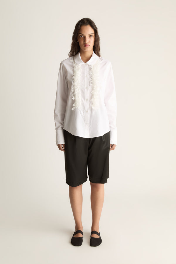 Front view of a woman wearing a white long-sleeve shirt with ruffle trim and black knee-length shorts against a white studio background