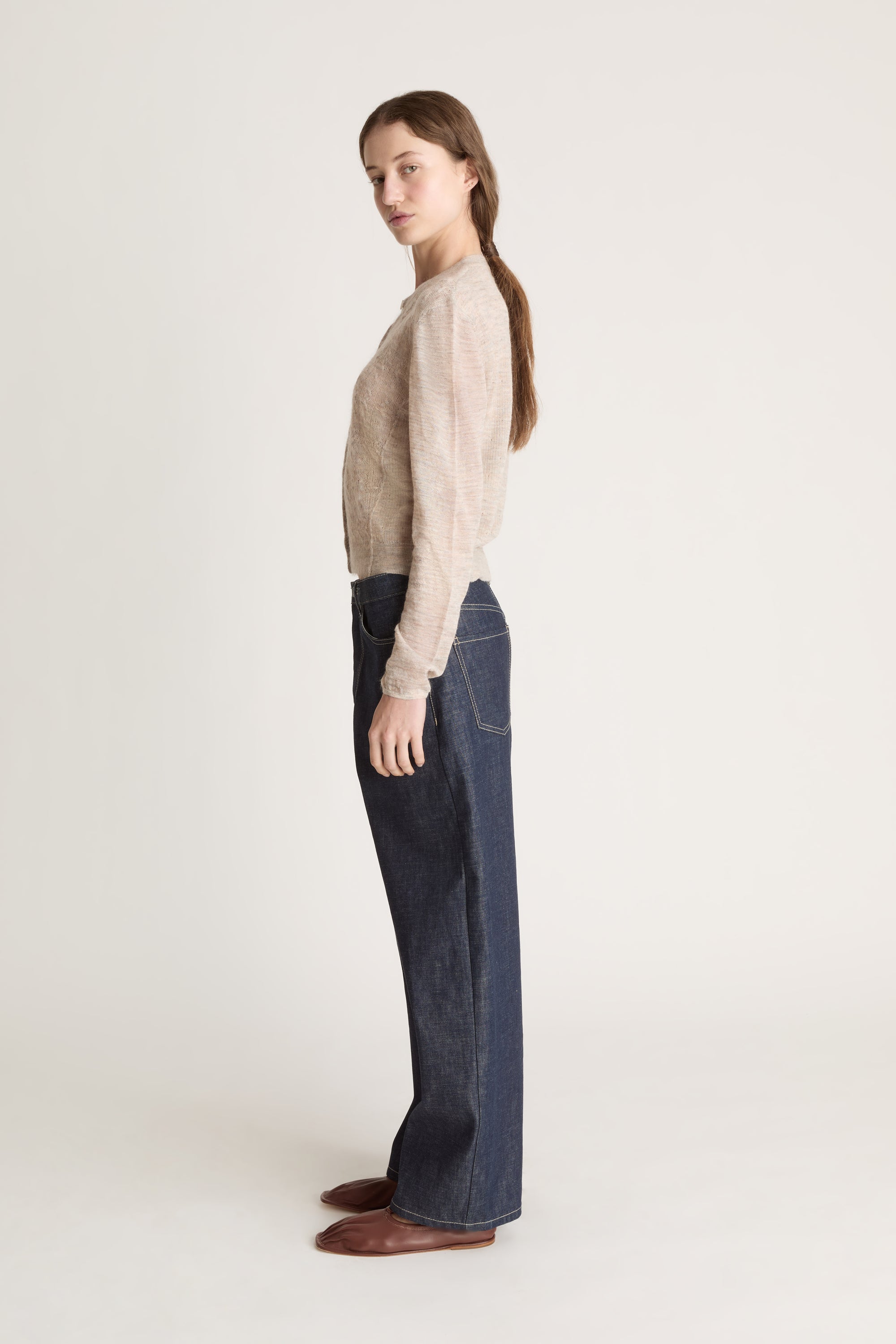 Person wearing a beige cardigan and dark jeans on a white background
