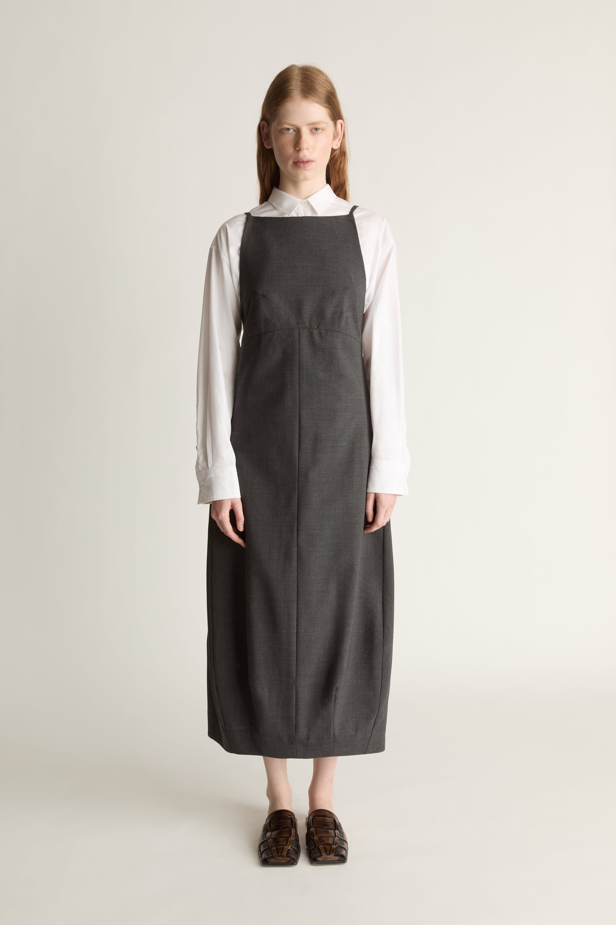 Person wearing a long dark apron style dress over a light shirt on a plain background