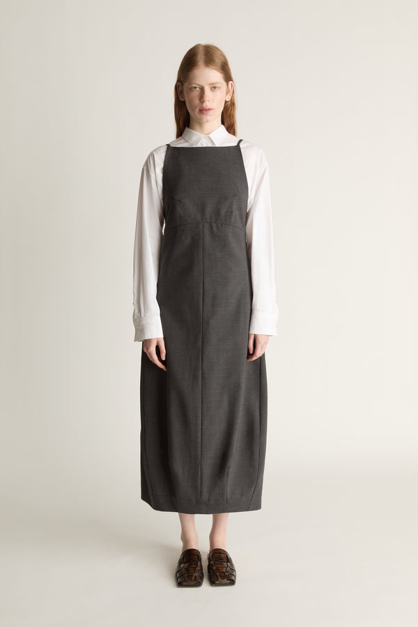 Person wearing a long dark apron style dress over a light shirt on a plain background